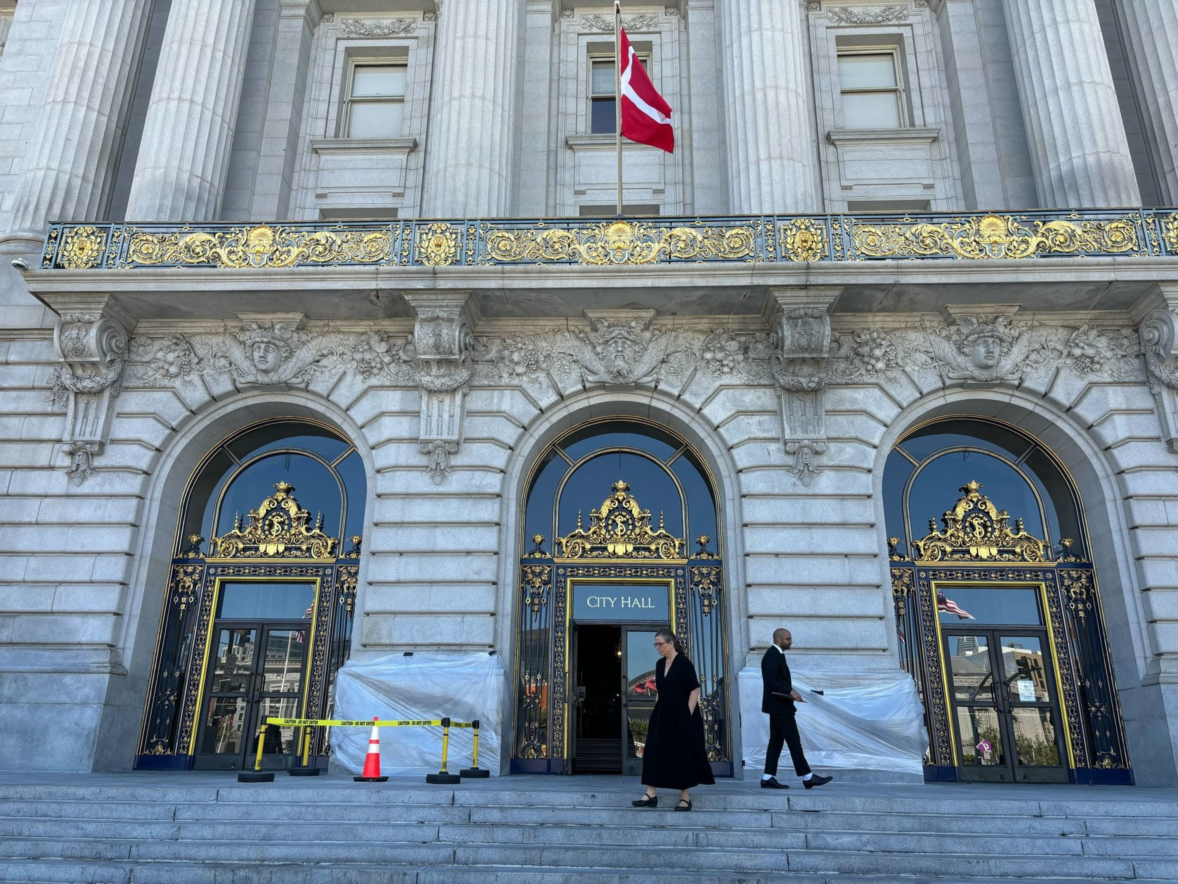 The image shows an ornate building facade with tall columns and gold detailing on arched doors. &quot;City Hall&quot; is written above the center door. Two people walk past.