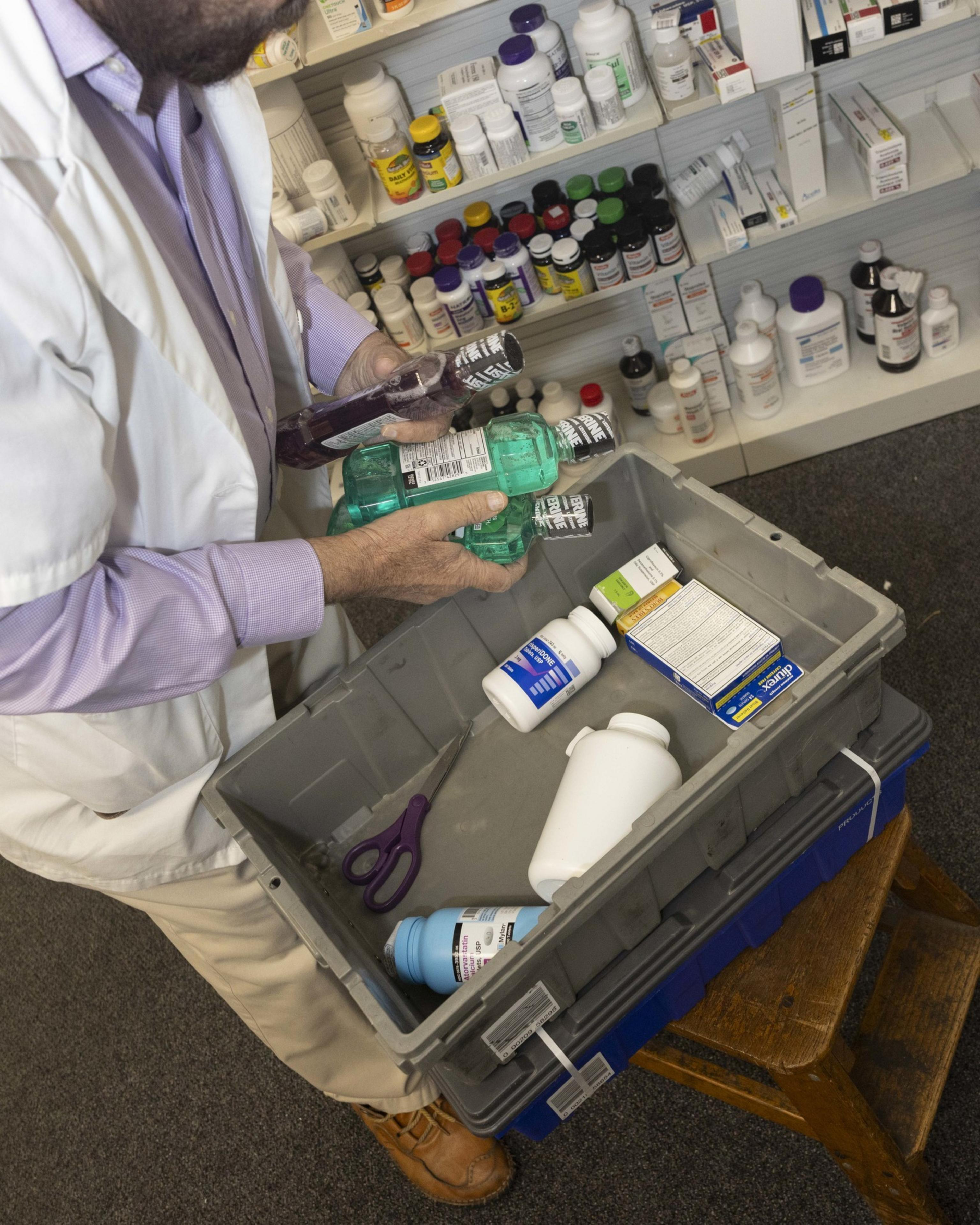 A person in a white lab coat is holding bottles of mouthwash above a bin with medicine bottles, scissors, and boxes. Shelves of more medicine are in the background.