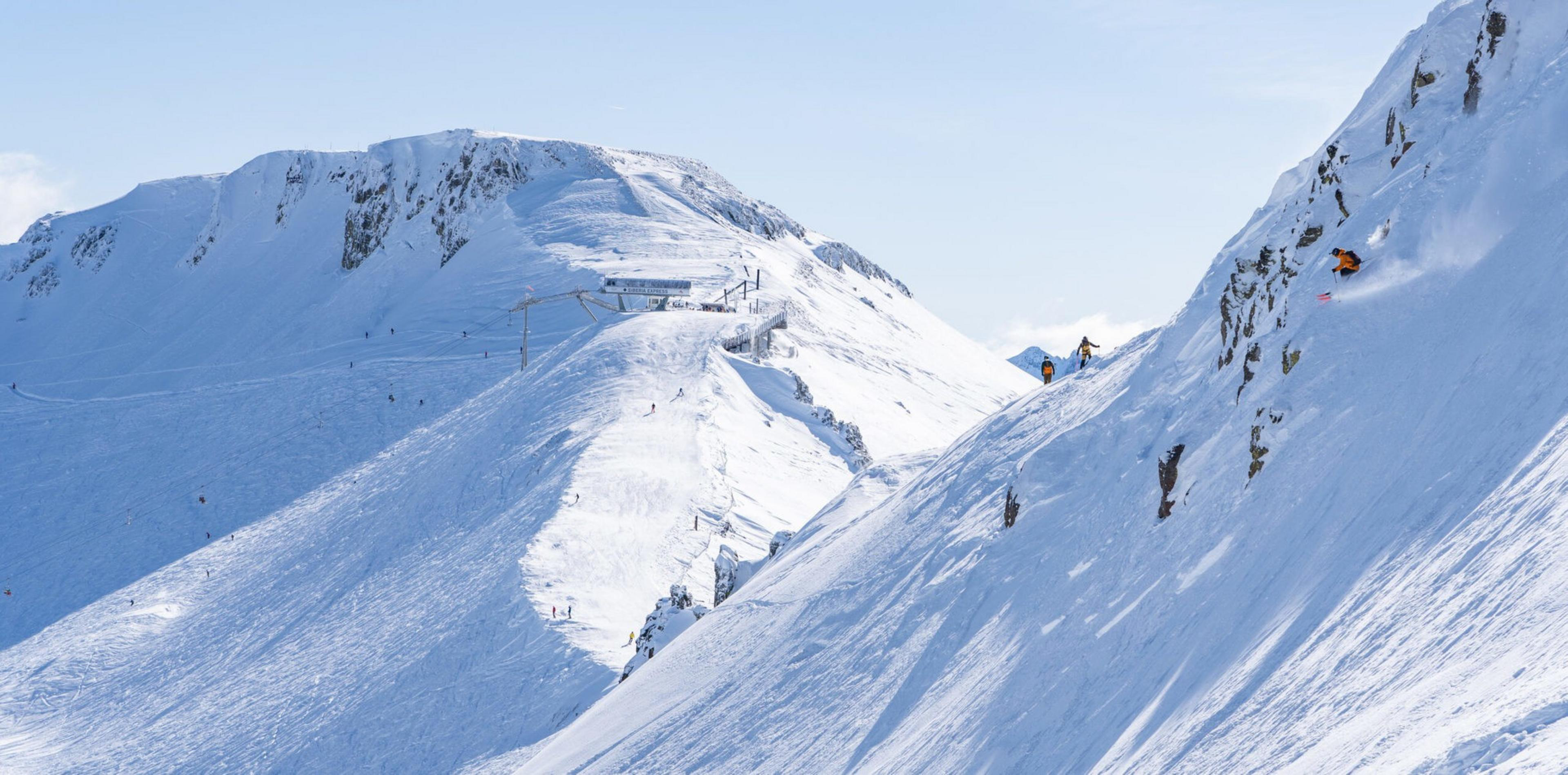 Snowboarders and skiers are descending steep, snowy mountain slopes with rocky patches under a clear blue sky.