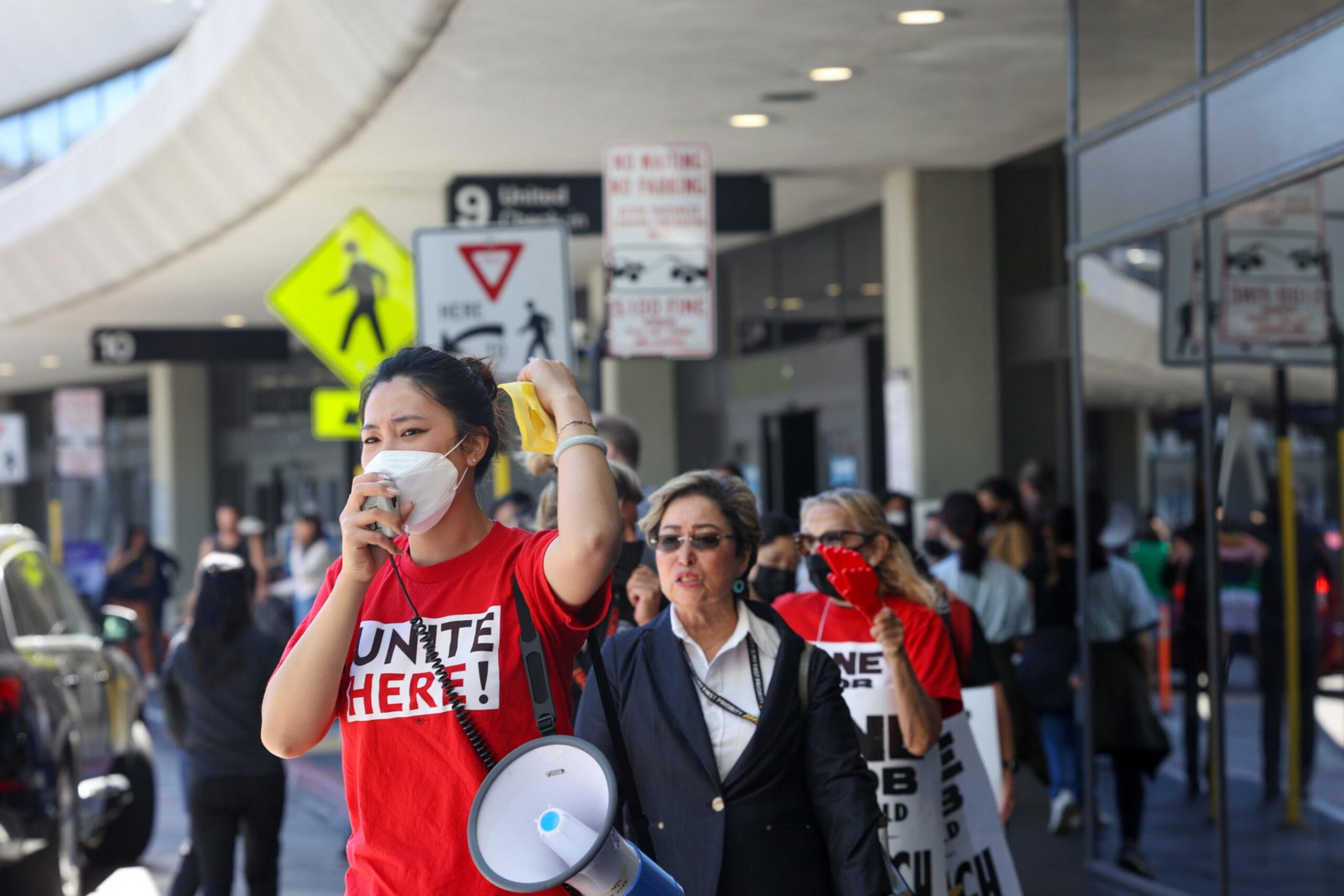 A woman in a red "UNITE HERE!" shirt speaks into a megaphone while a crowd, including another person in red, walk behind her near a building entrance.
