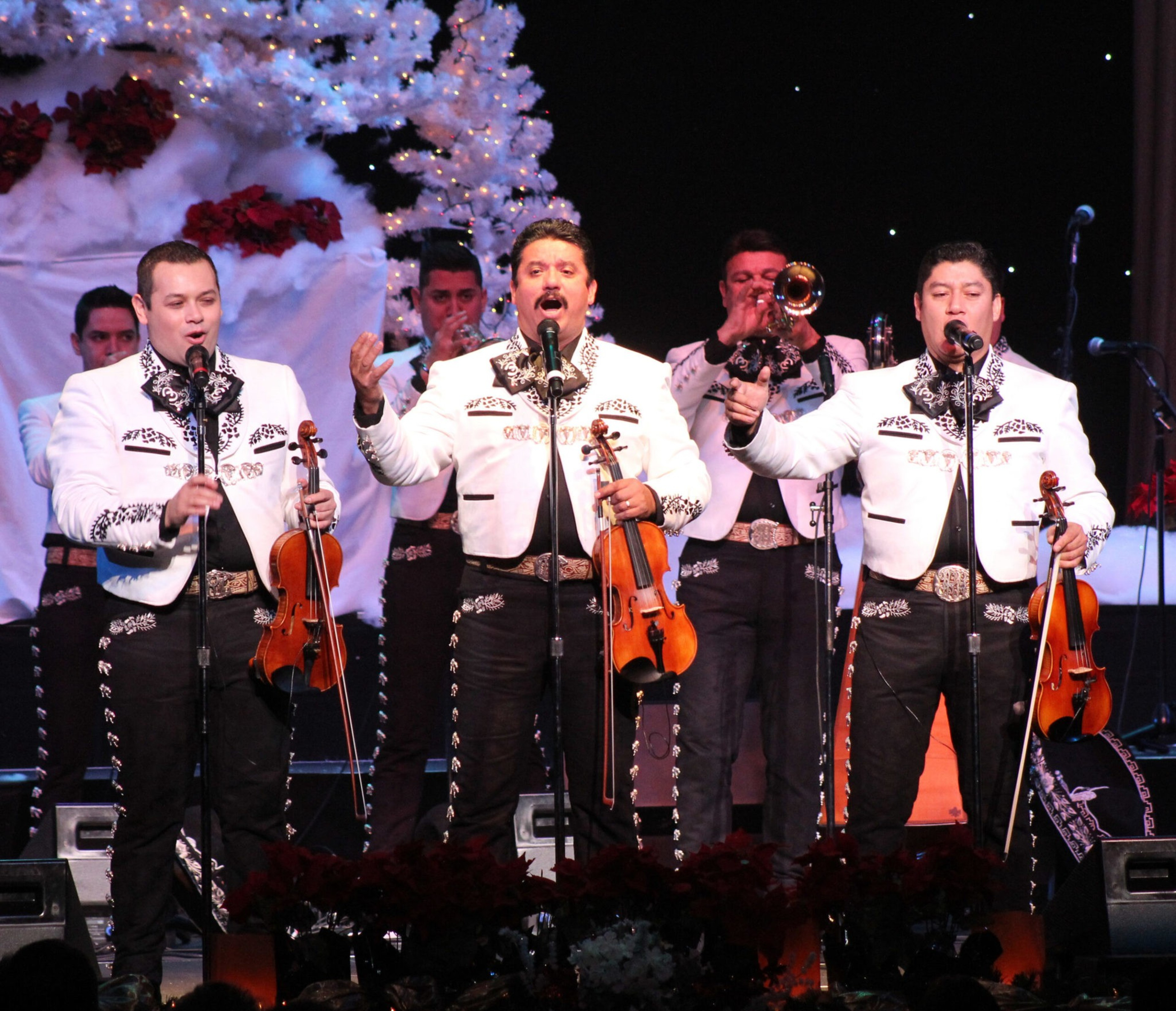 Three musicians in matching white charro jackets and black pants perform on stage, holding violins and singing, with trumpet players visible in the background.