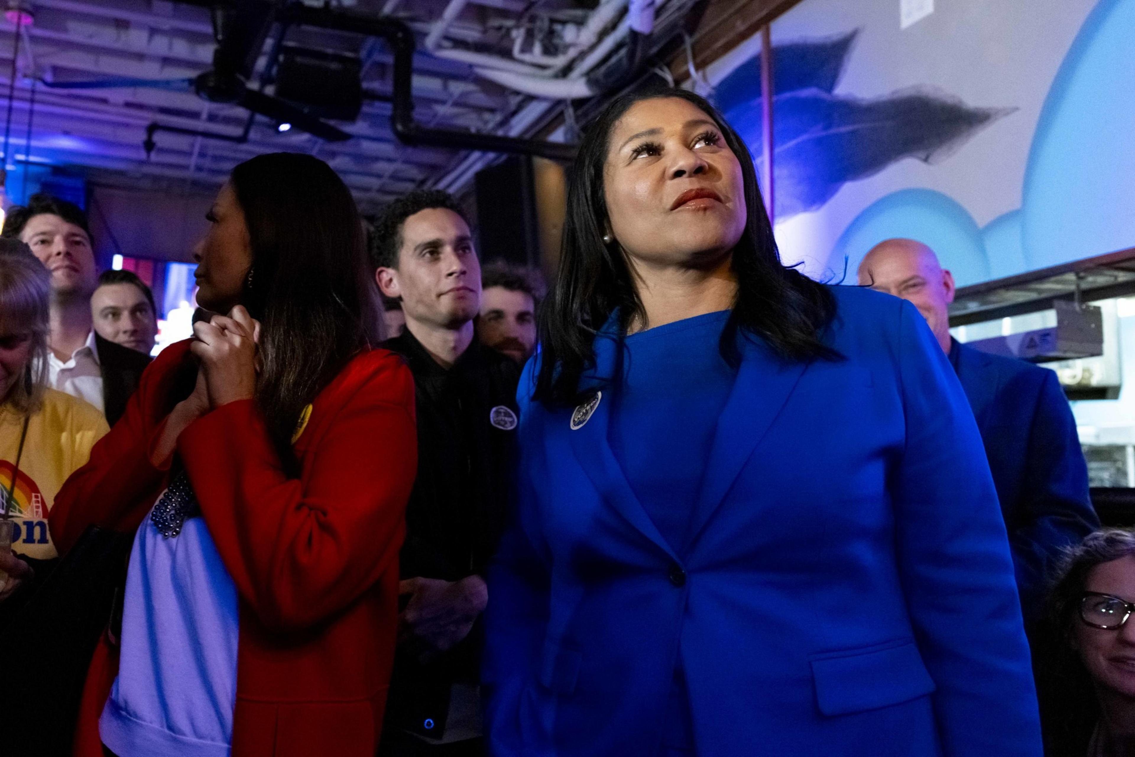A woman in a blue blazer stands confidently under colorful lighting, surrounded by a group of people, some in casual attire, in a dimly lit room.