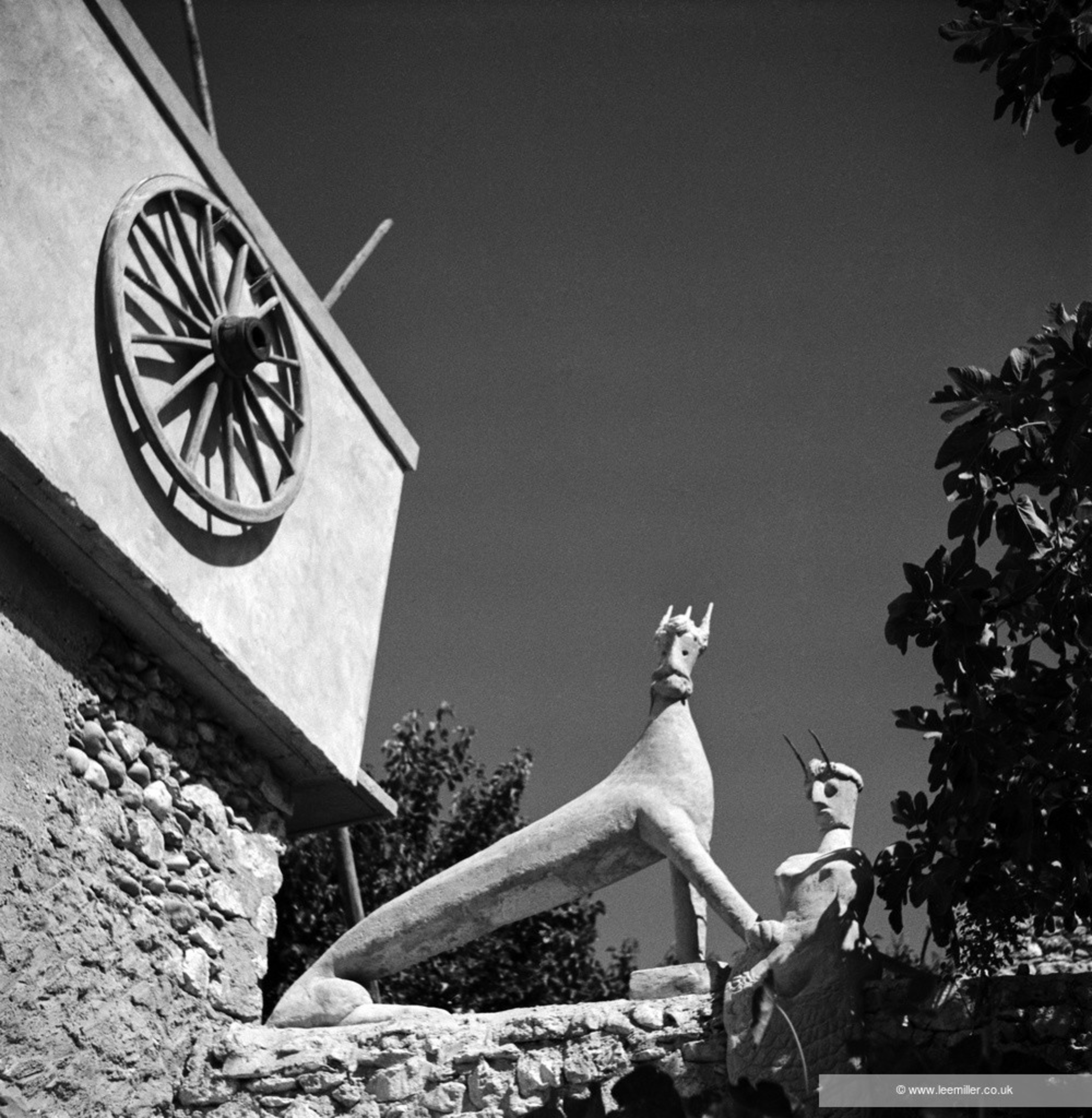 The image shows two abstract sculptures resembling animals with elongated bodies and horns, positioned on a stone wall. A wooden wagon wheel decorates a nearby building wall.