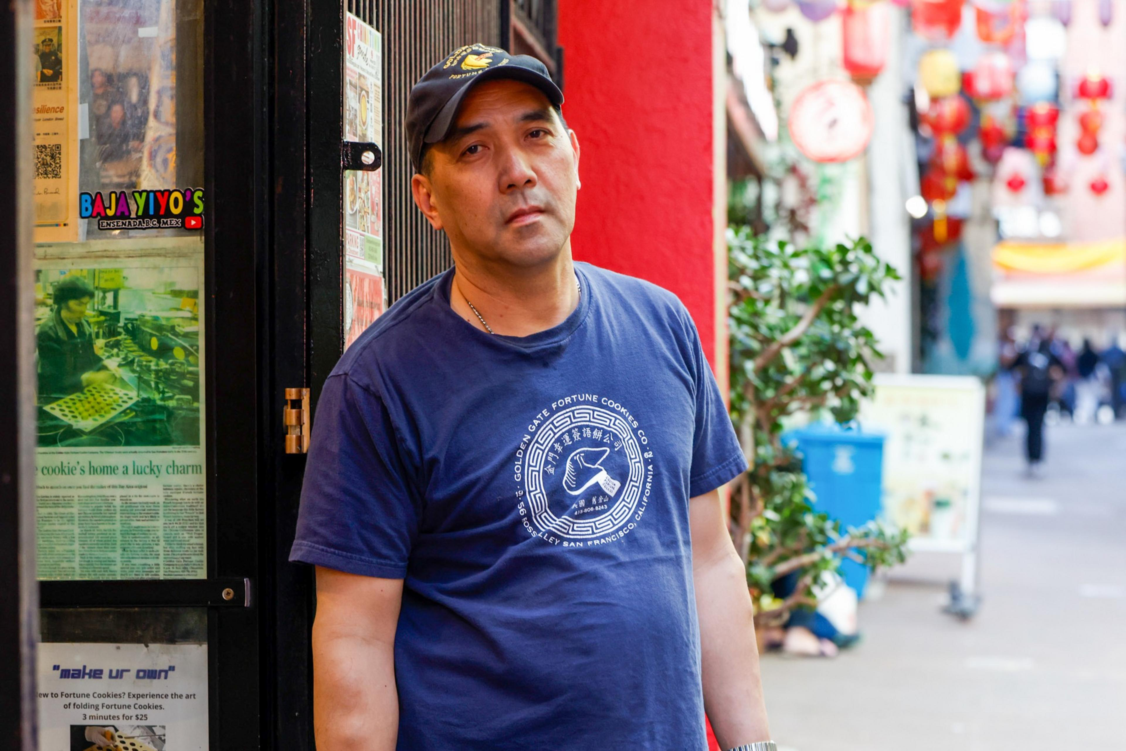 A man in a navy shirt with a printed design leans against a red wall in a colorful street decorated with lanterns. He appears relaxed and thoughtful.
