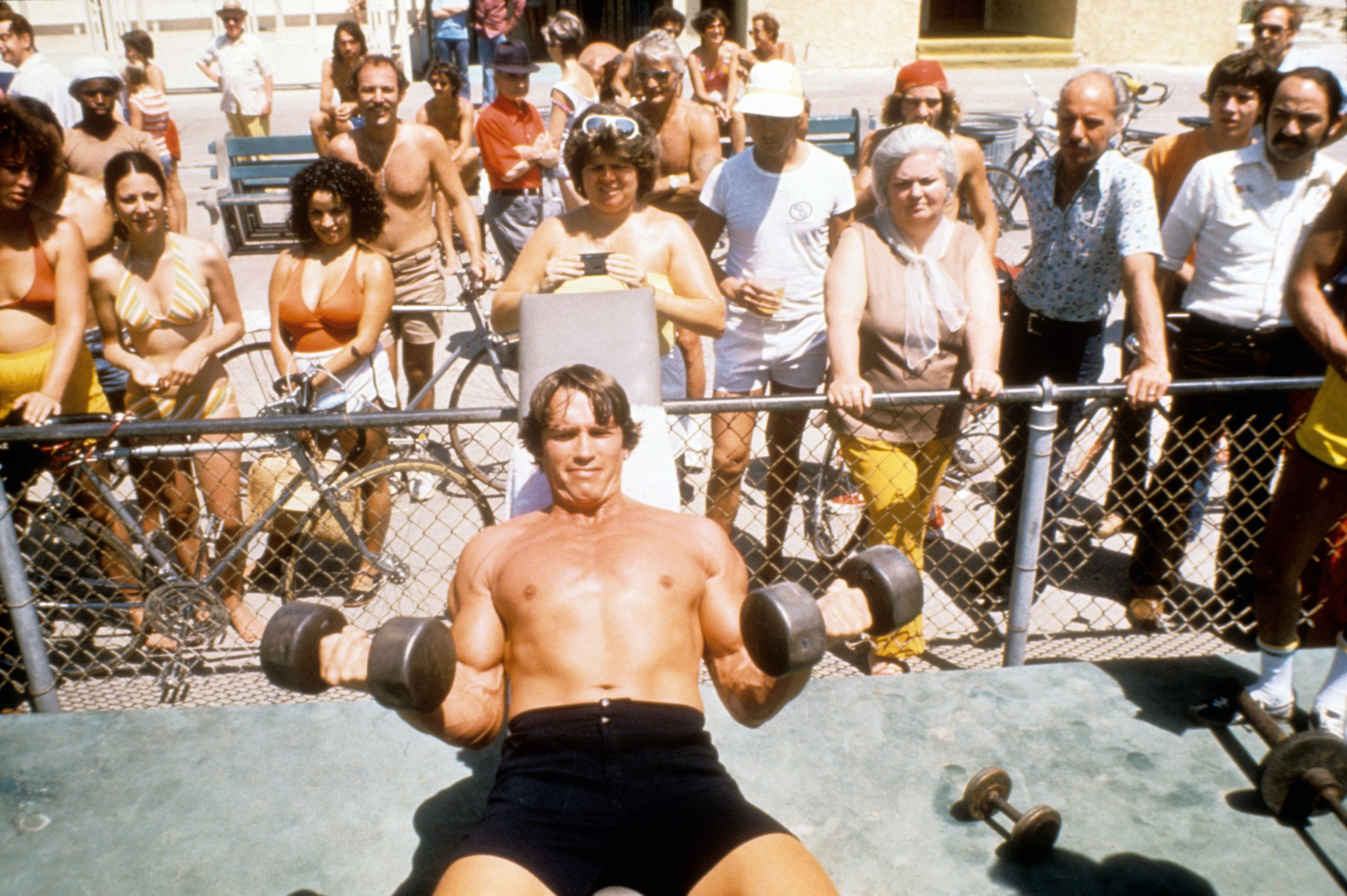 A muscular man lifts dumbbells on a bench while a diverse group of shirtless and casually dressed people watch behind a chain-link fence.