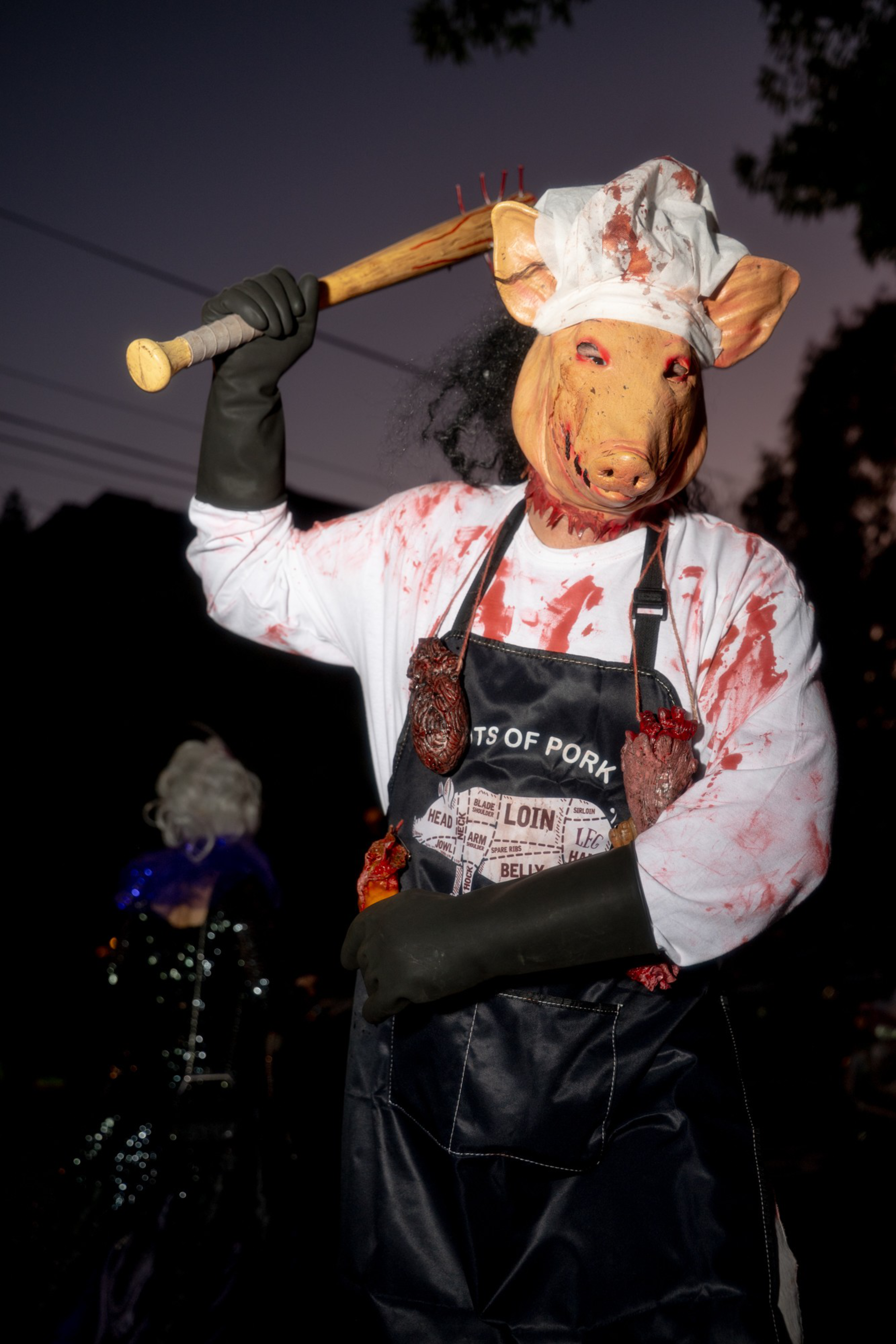A person wears a bloody pig mask and chef's hat, a bloodstained apron with meat cuts, black gloves, and wields a wooden bat with nails.