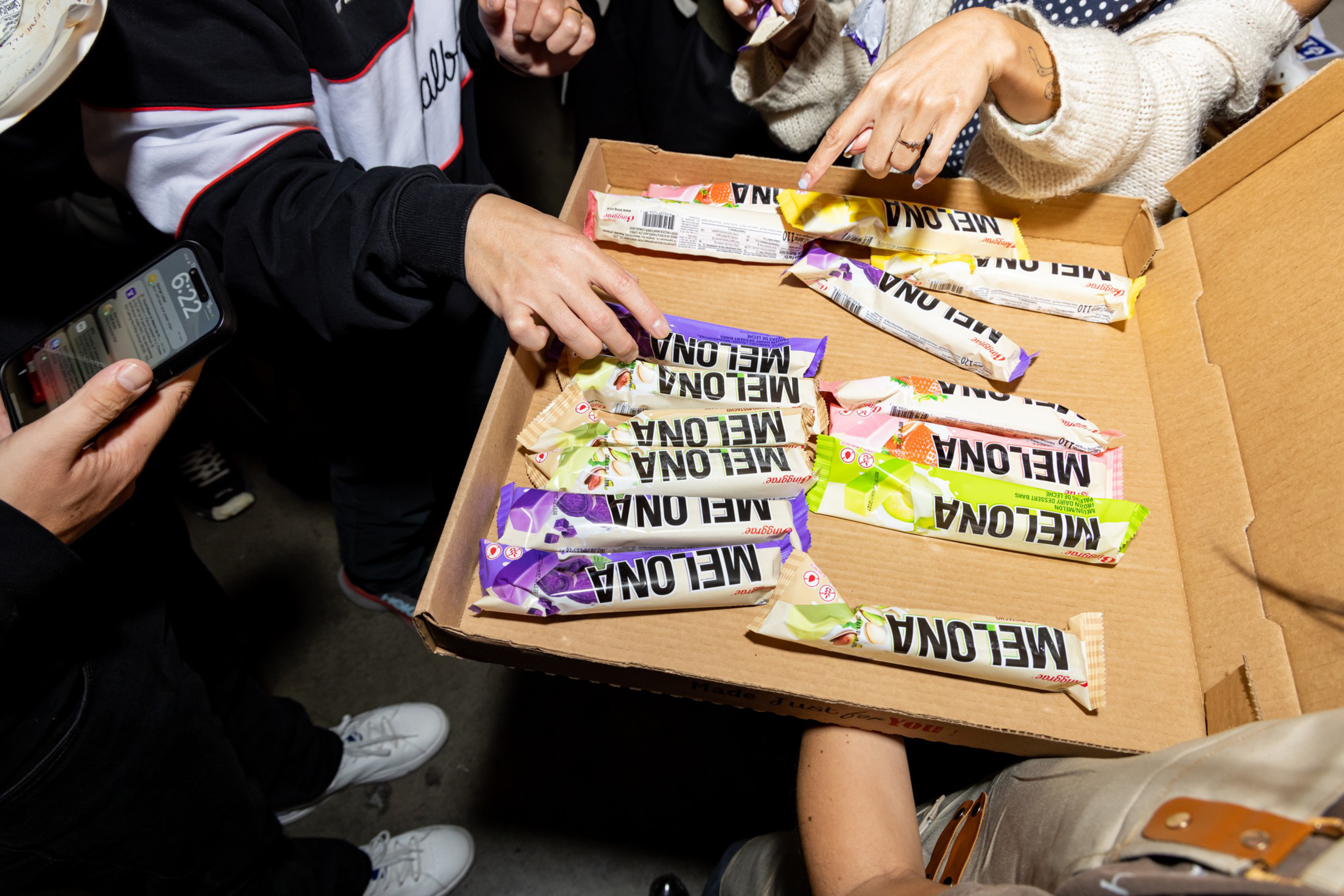 A group of people reaching into a pizza box filled with various colorful Melona ice cream bars.