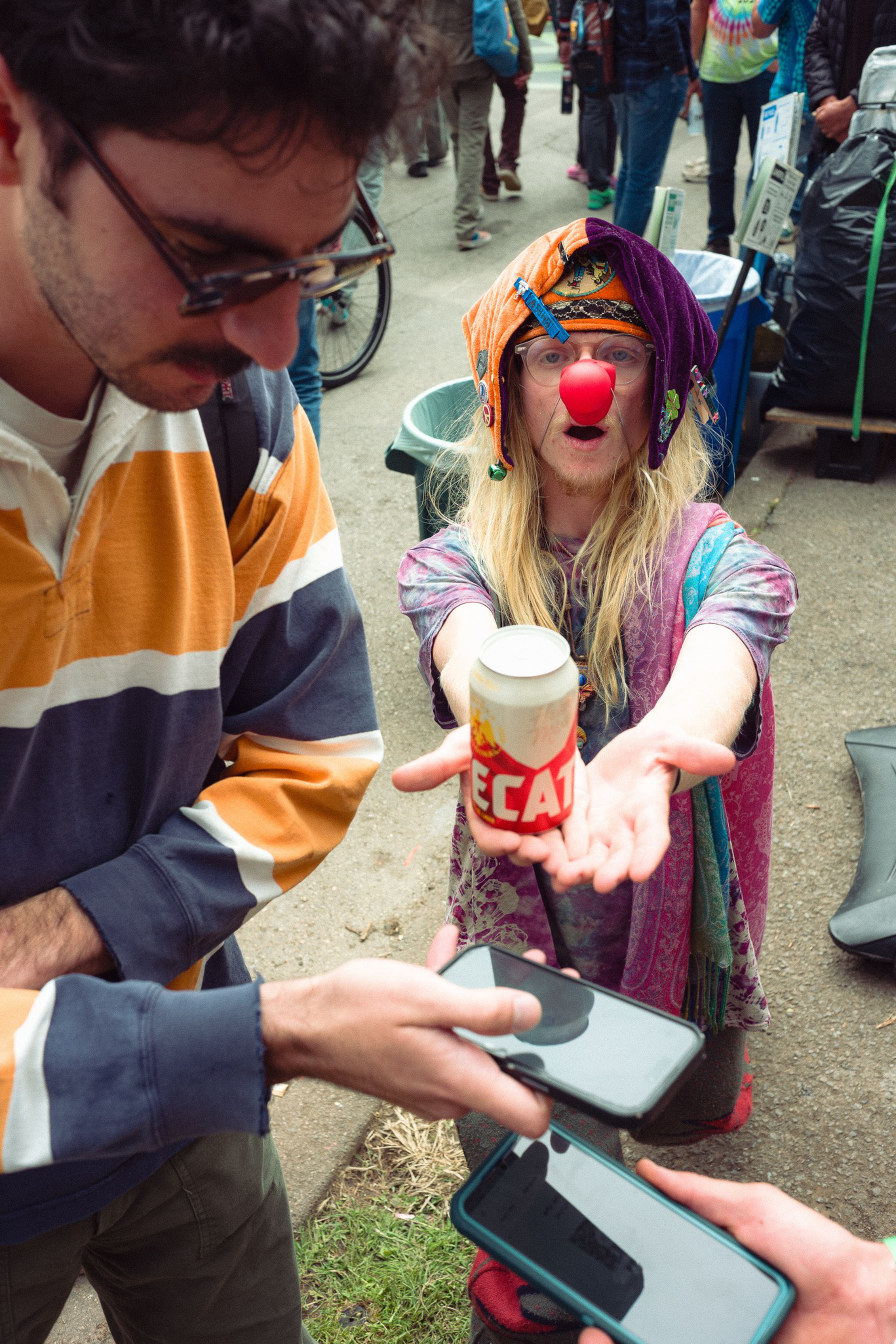 A person with a colorful jester hat, red clown nose, and long blonde hair holds out a beer can, while two people nearby look at their phones.