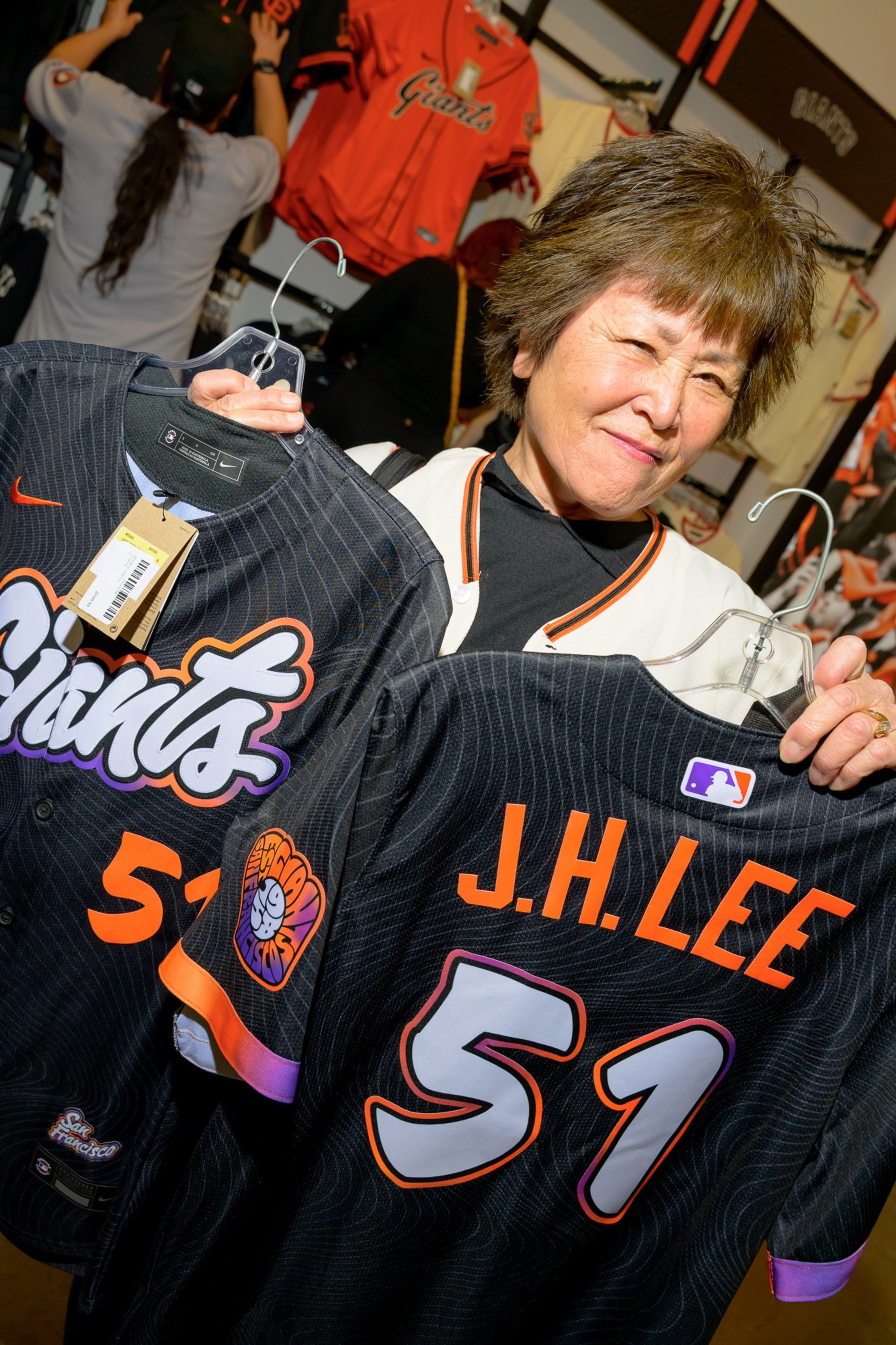 A person holds two black Giants baseball jerseys with the number 51. One jersey displays &quot;J.H. Lee&quot; on the back. The jerseys are hung on hangers.