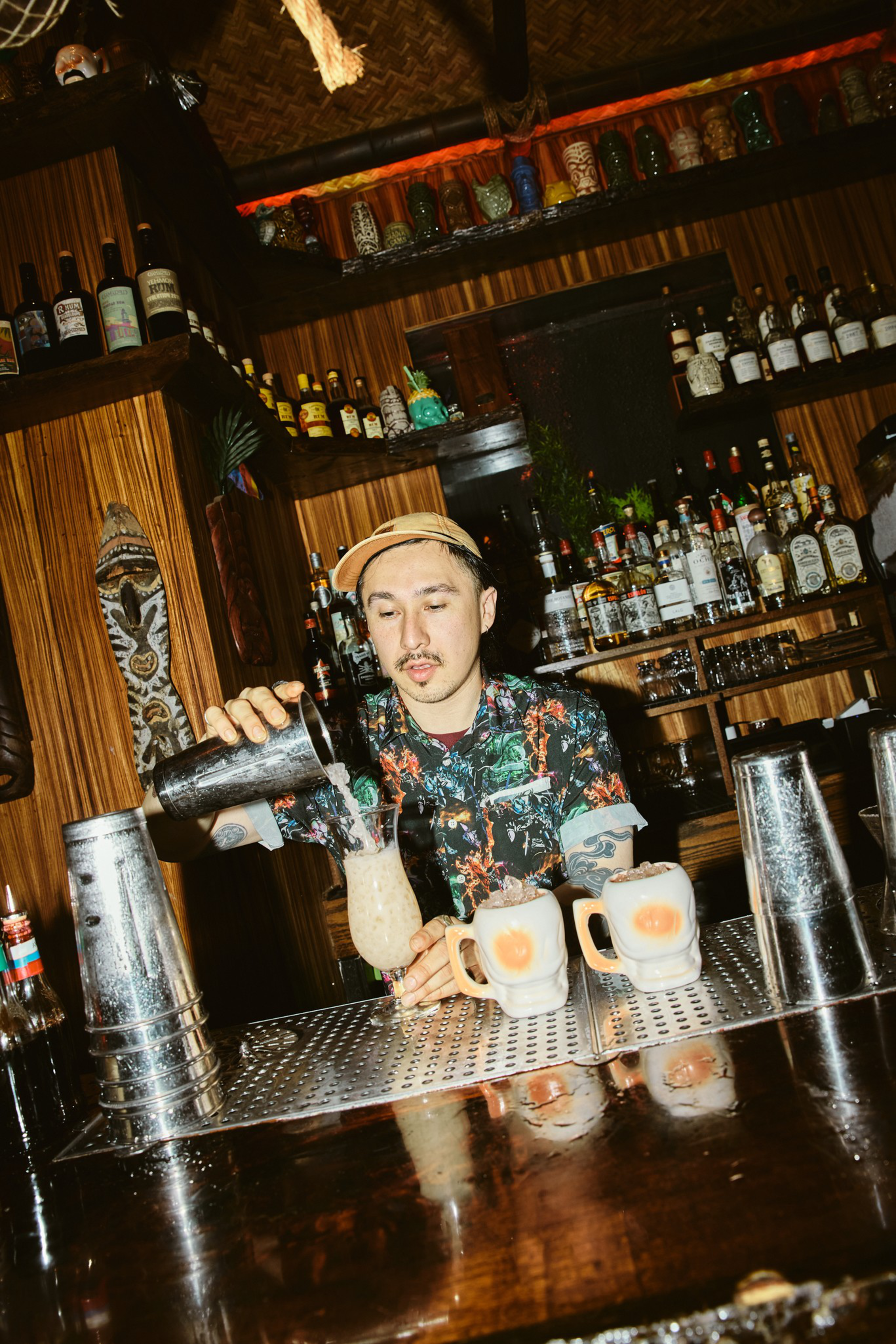A bartender wearing a floral shirt and cap pours a drink from a shaker into a tiki glass, with two frothy mugs and metal shakers on the bar.
