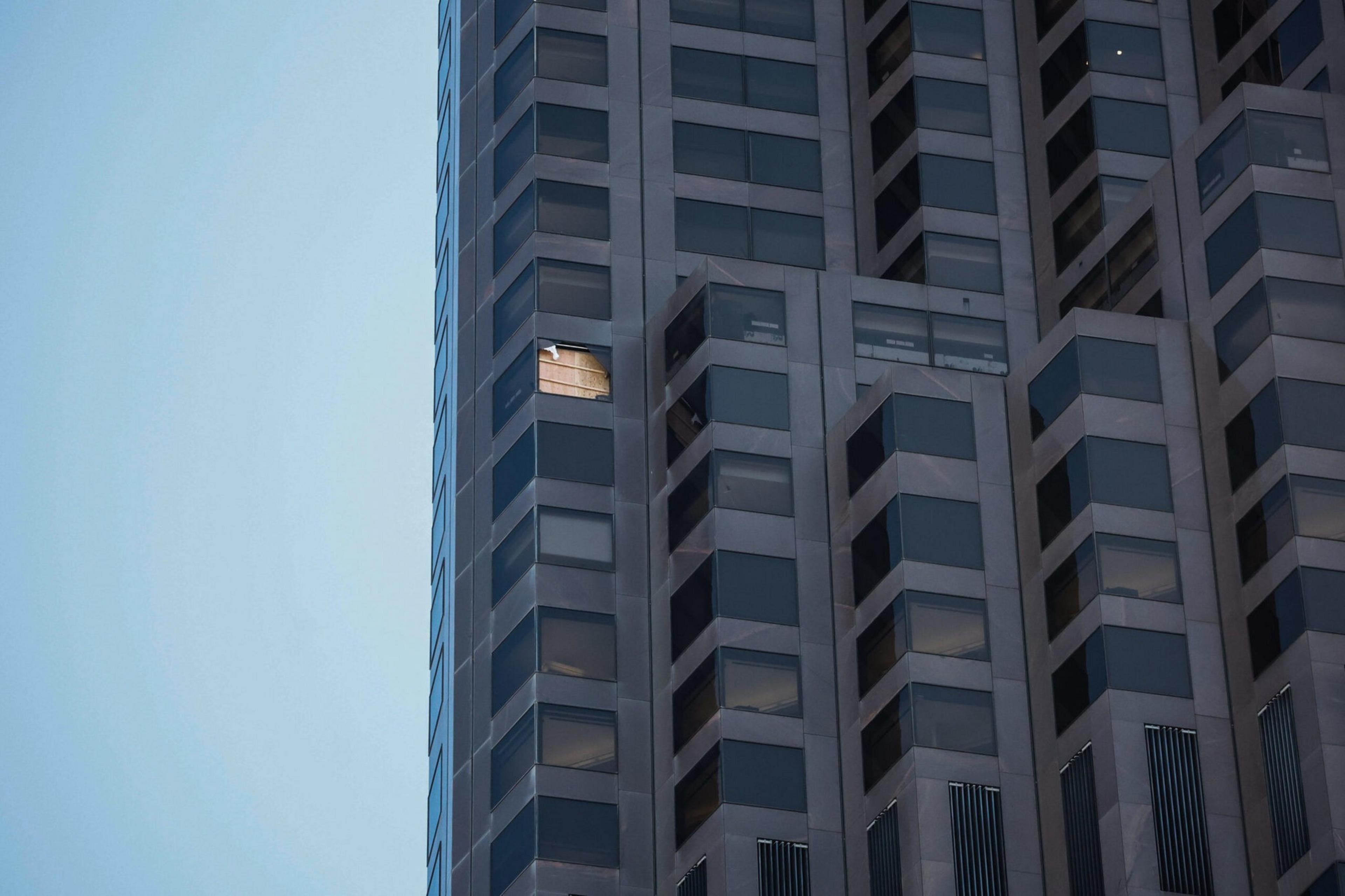 A tall building with many dark windows has one window open, revealing a warm light from inside against a clear blue sky.