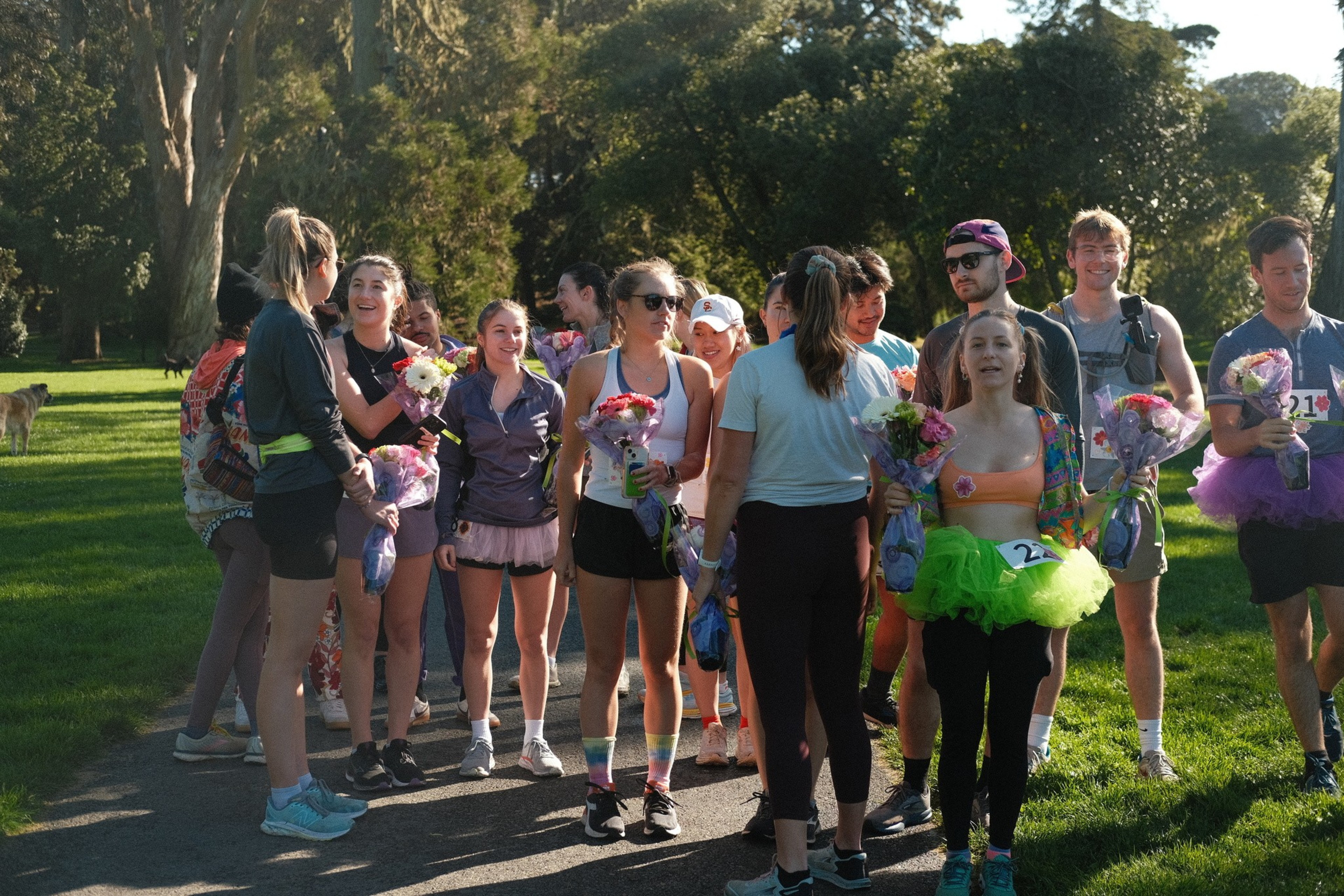 A group of people in athletic attire stands on a path in a sunny park, holding bouquets of flowers. Some are wearing tutus, suggesting a fun or themed event.