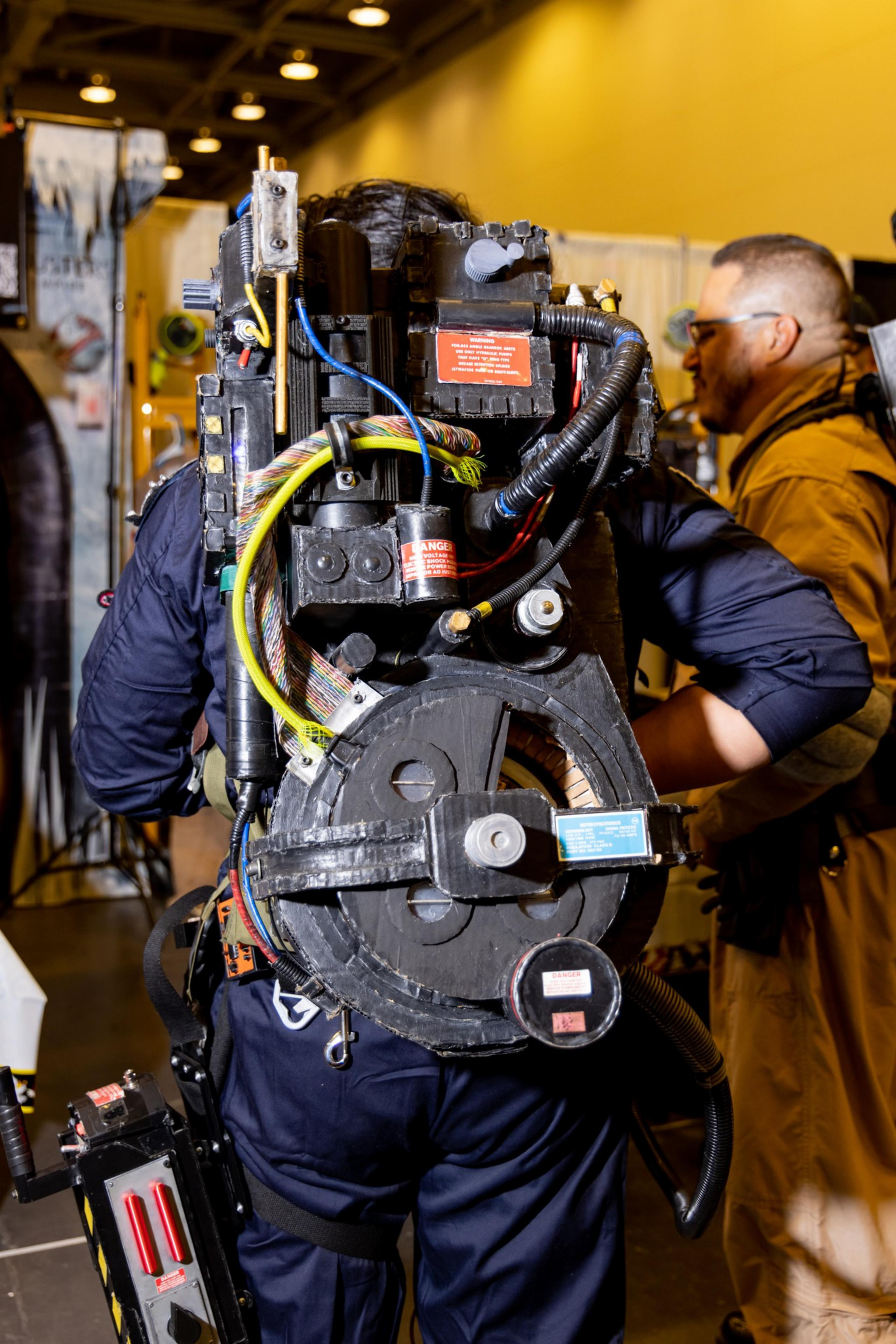 A person is wearing a detailed, elaborate costume resembling a Ghostbuster's proton pack, filled with wires and gadgets, in a well-lit indoor setting.