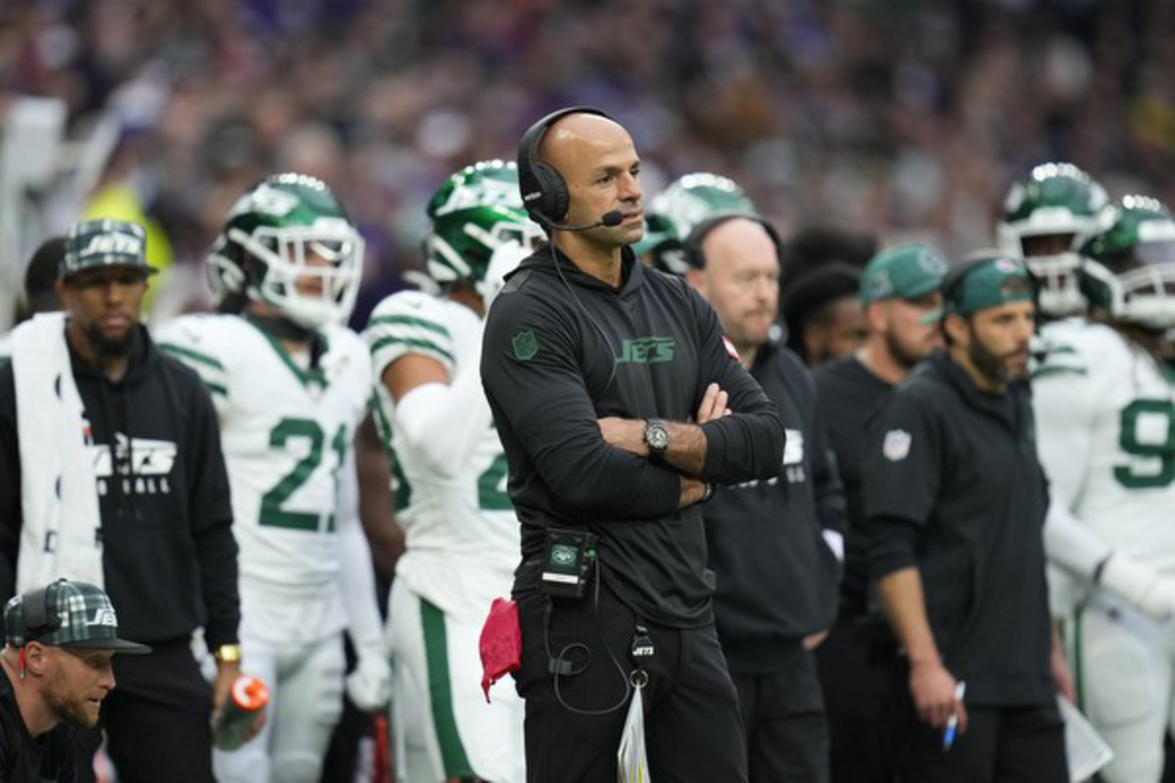 A football coach wearing a headset stands on the sideline with arms crossed. Players in green and white uniforms, also stand in the background.