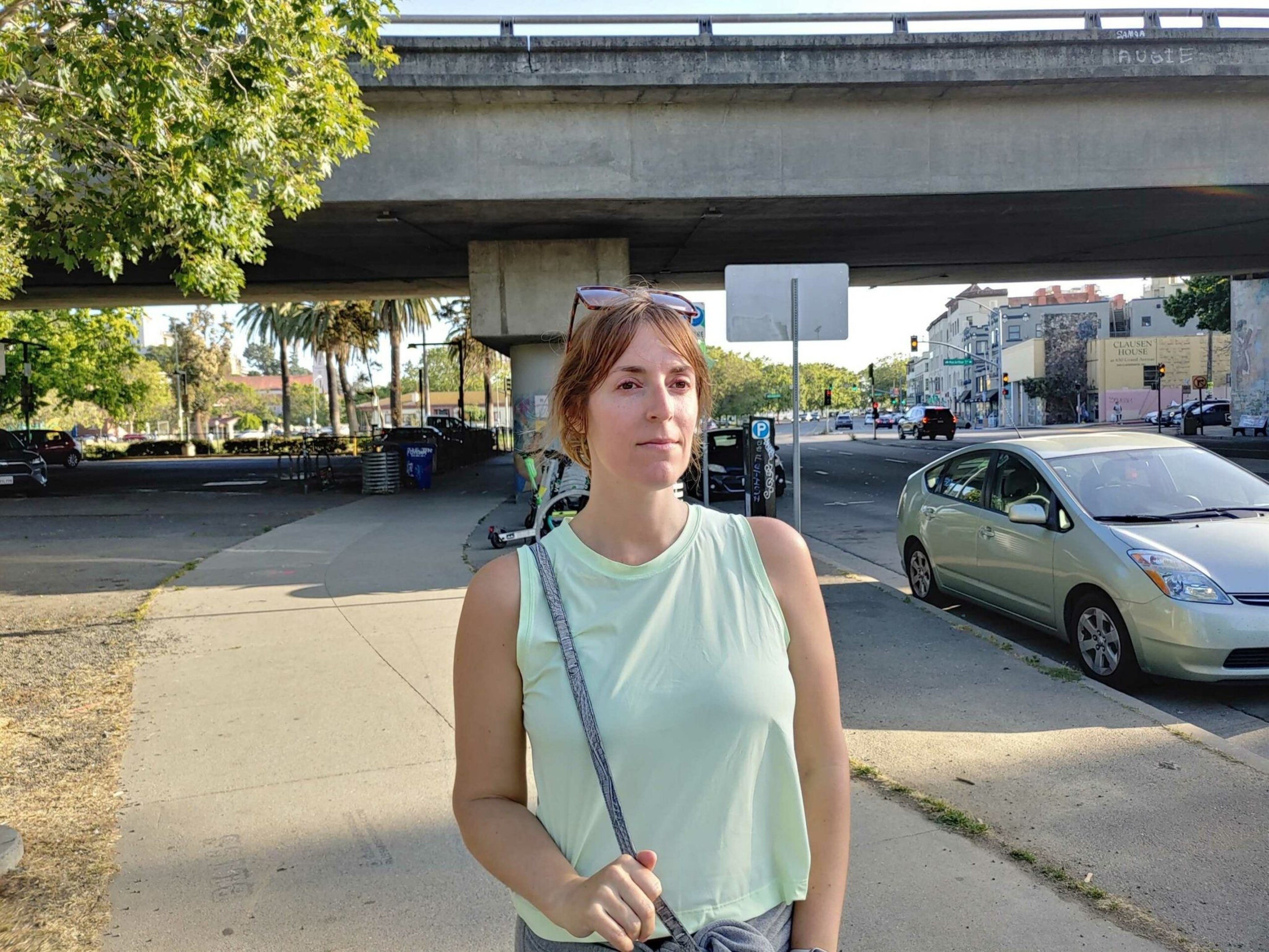 A woman with sunglasses on her head stands on a sunny sidewalk near a parked light green car under a concrete overpass.