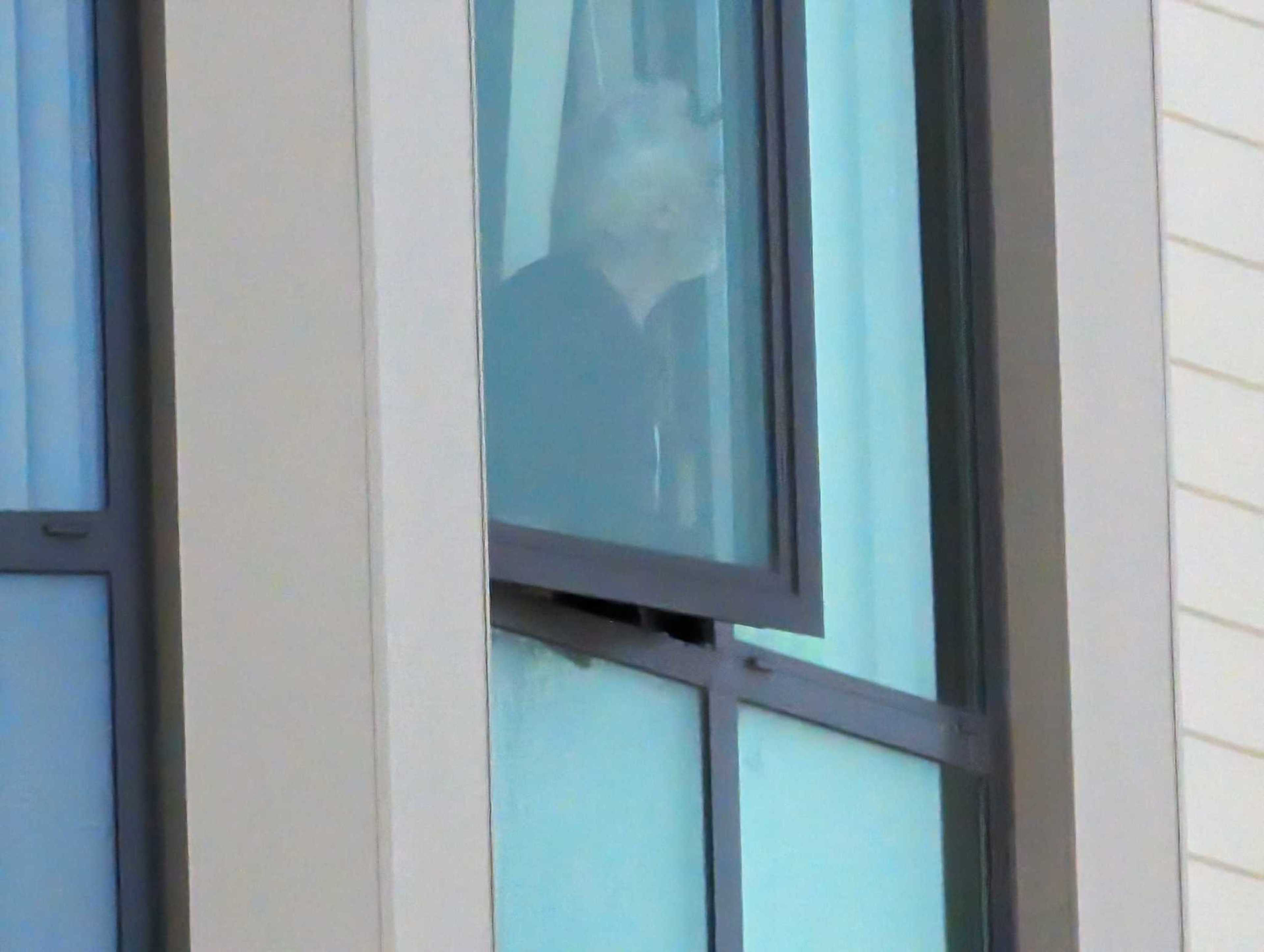 A person with light hair is partially visible through a slightly open window, looking outside. The window has a beige frame.