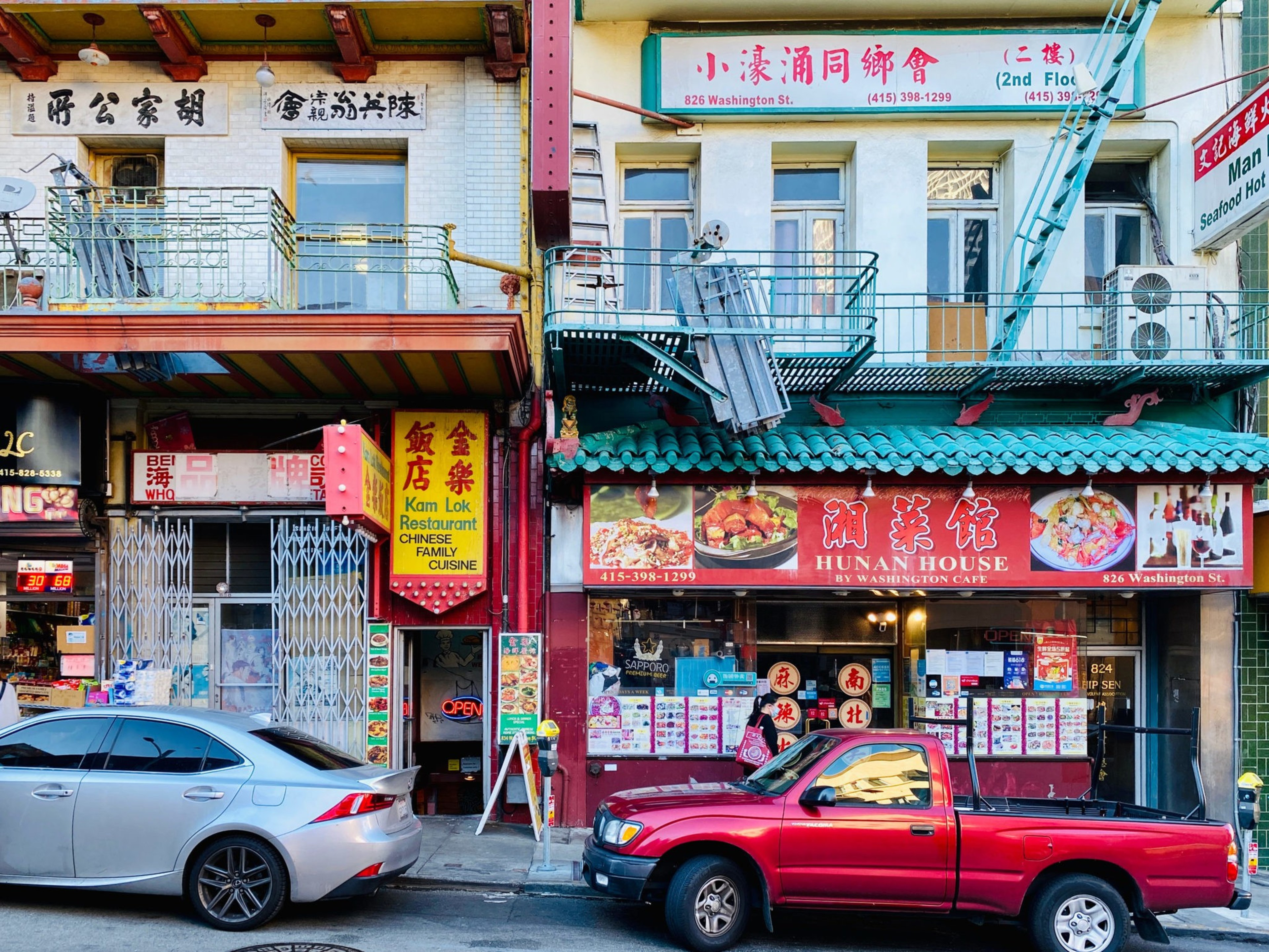 Two parked cars, a silver sedan and a red pickup, in front of Chinese restaurants with colorful signs and menus on a city street.