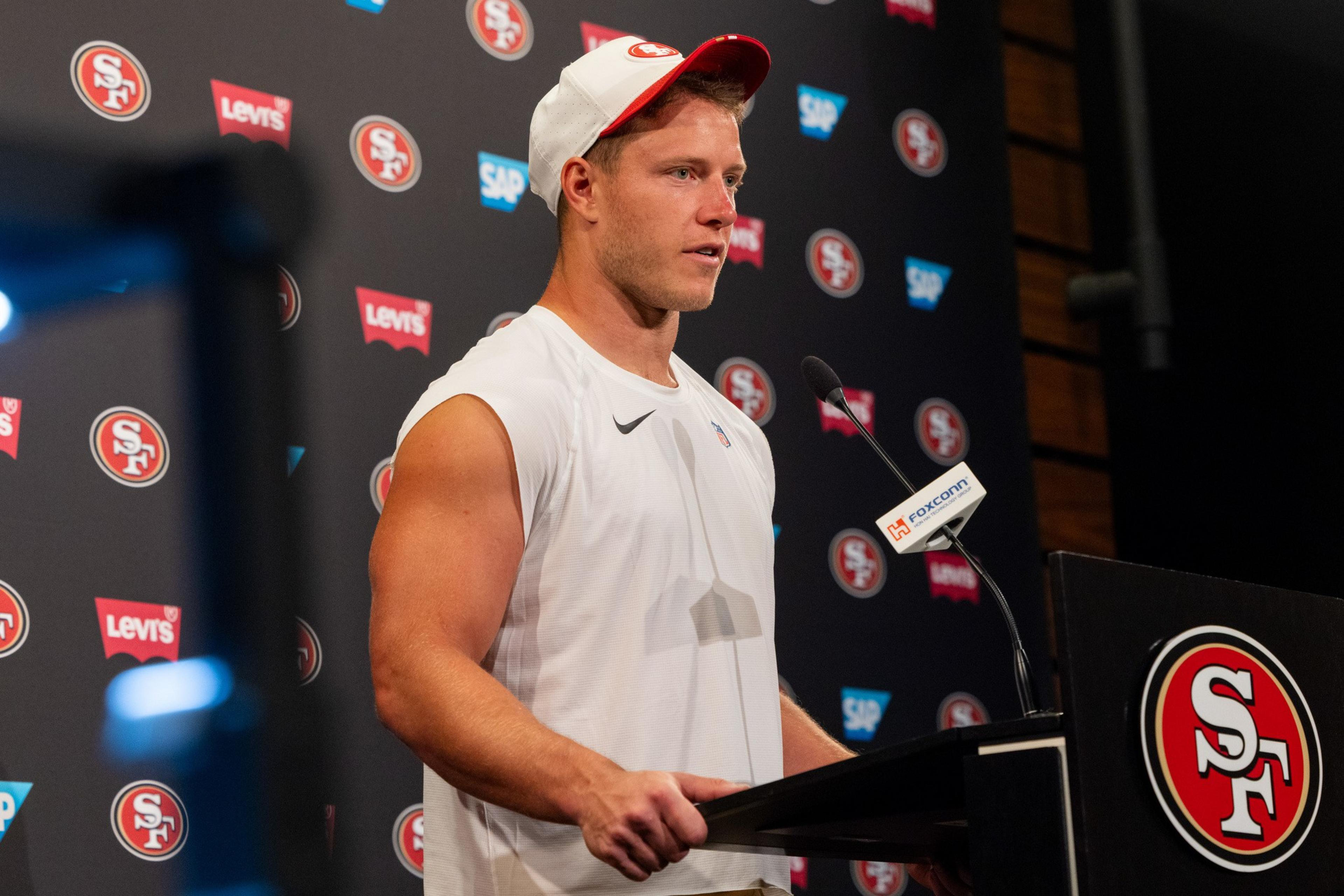 A man in a sleeveless white shirt and a white and red cap stands at a podium speaking, with a backdrop of San Francisco 49ers and sponsor logos.