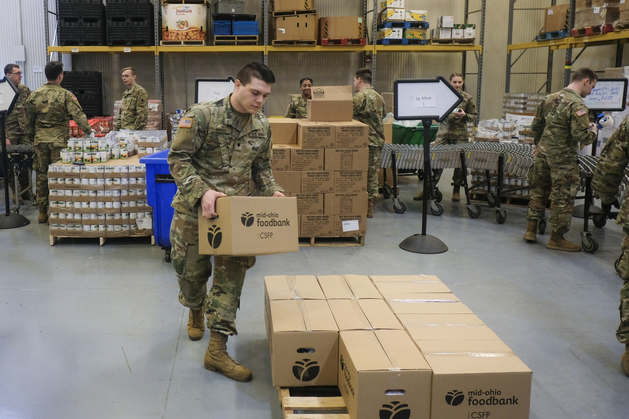 Soldiers in uniform are organizing and stacking boxes labeled "mid-ohio foodbank" inside a warehouse with shelves and supplies.
