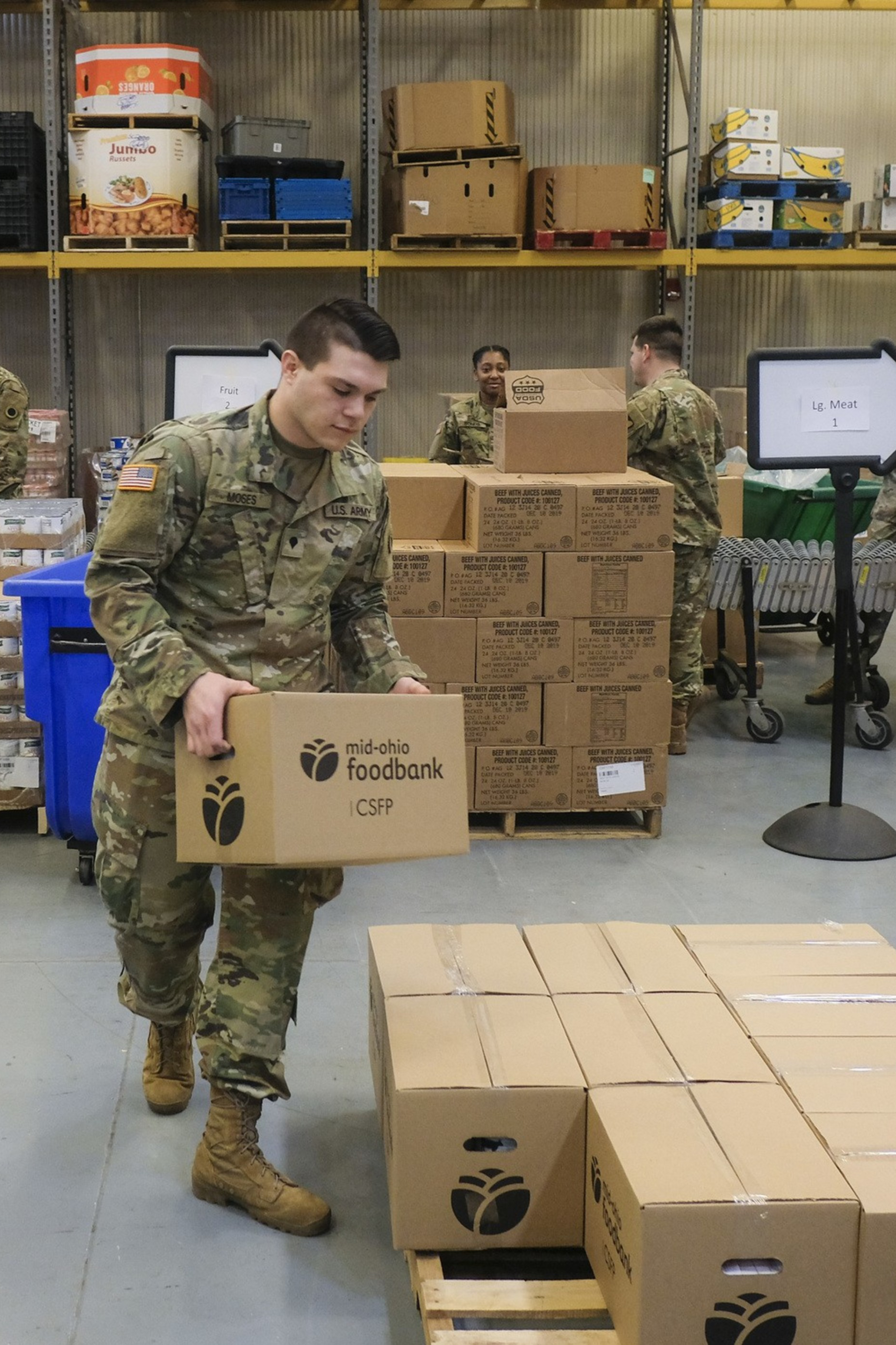 Soldiers in uniform are organizing and stacking boxes labeled "mid-ohio foodbank" inside a warehouse with shelves and supplies.