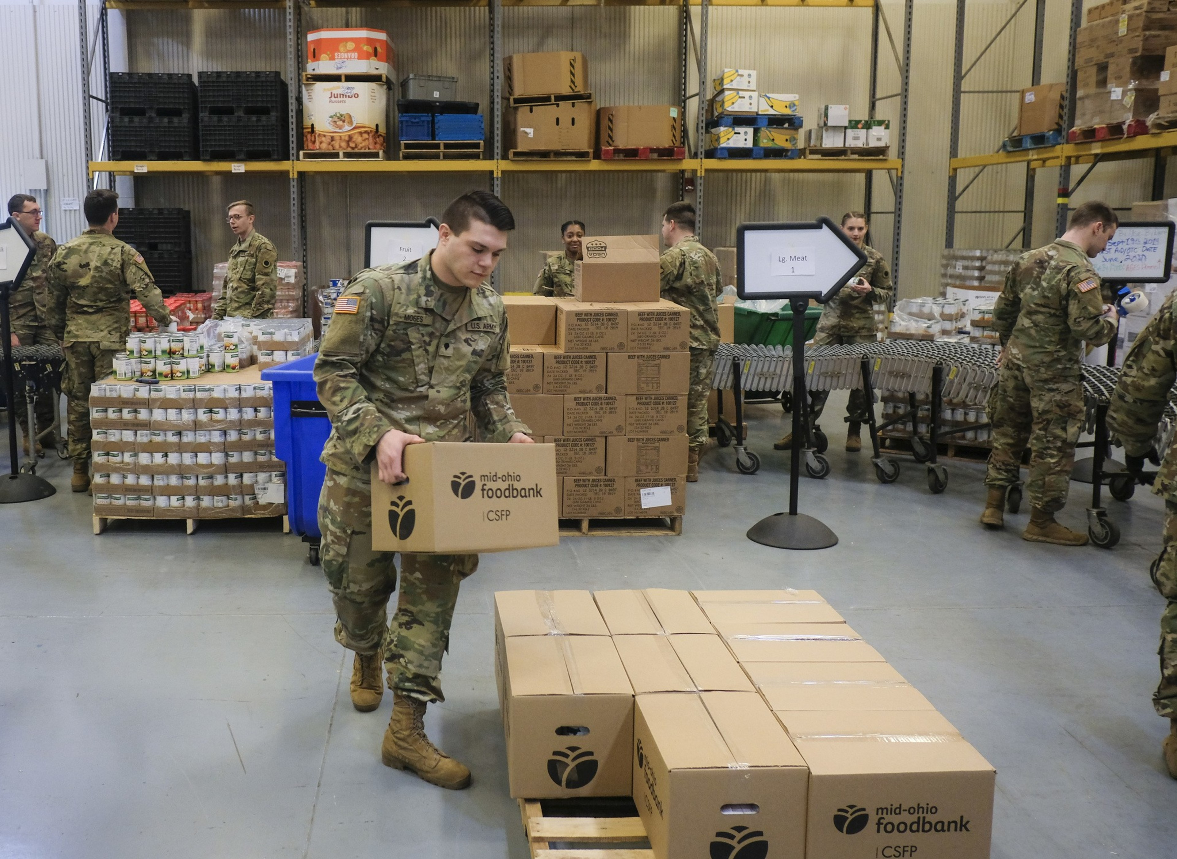 Soldiers in uniform are organizing and stacking boxes labeled "mid-ohio foodbank" inside a warehouse with shelves and supplies.