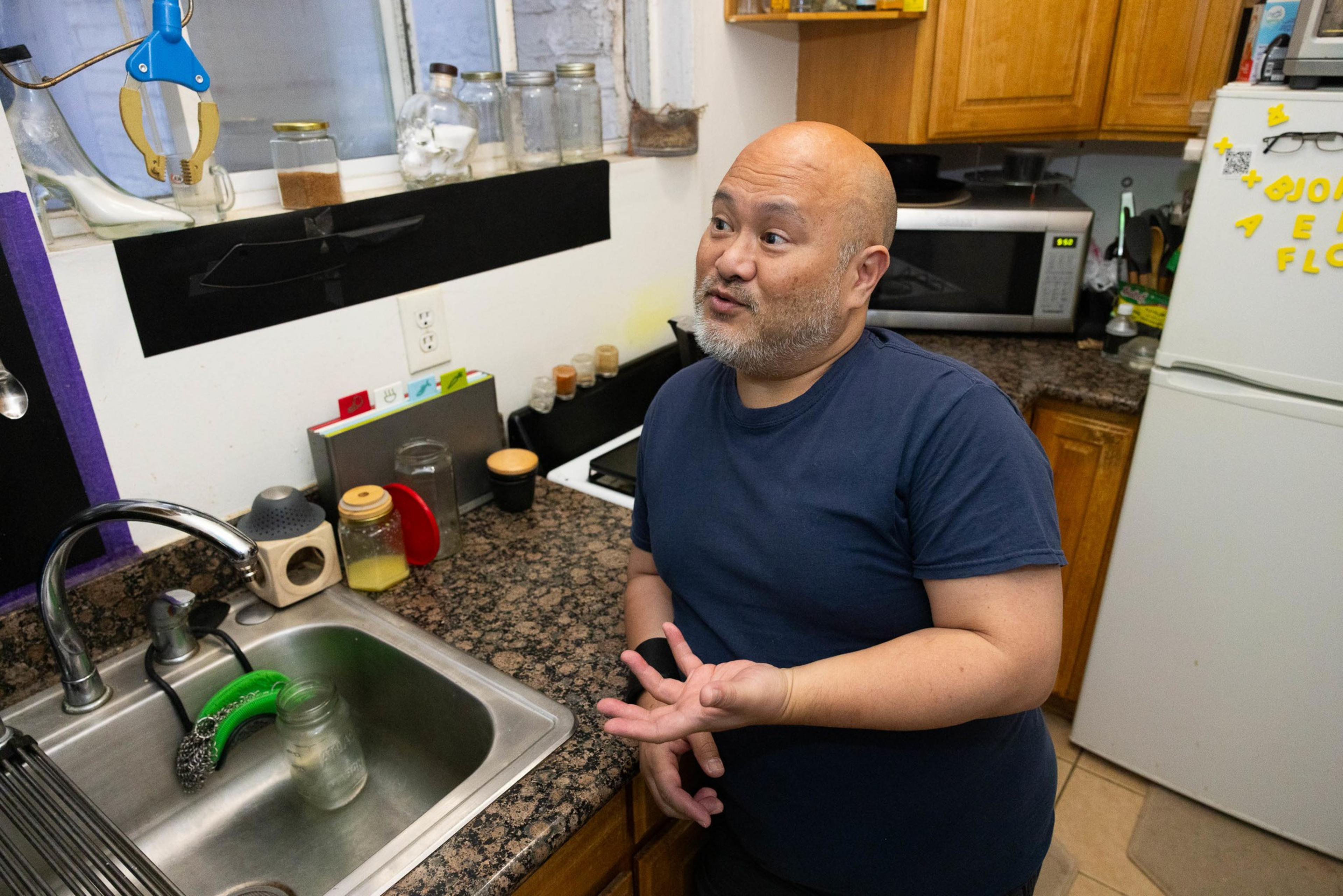 A man stands in a kitchen with utensils and jars, appearing to be in a conversation.