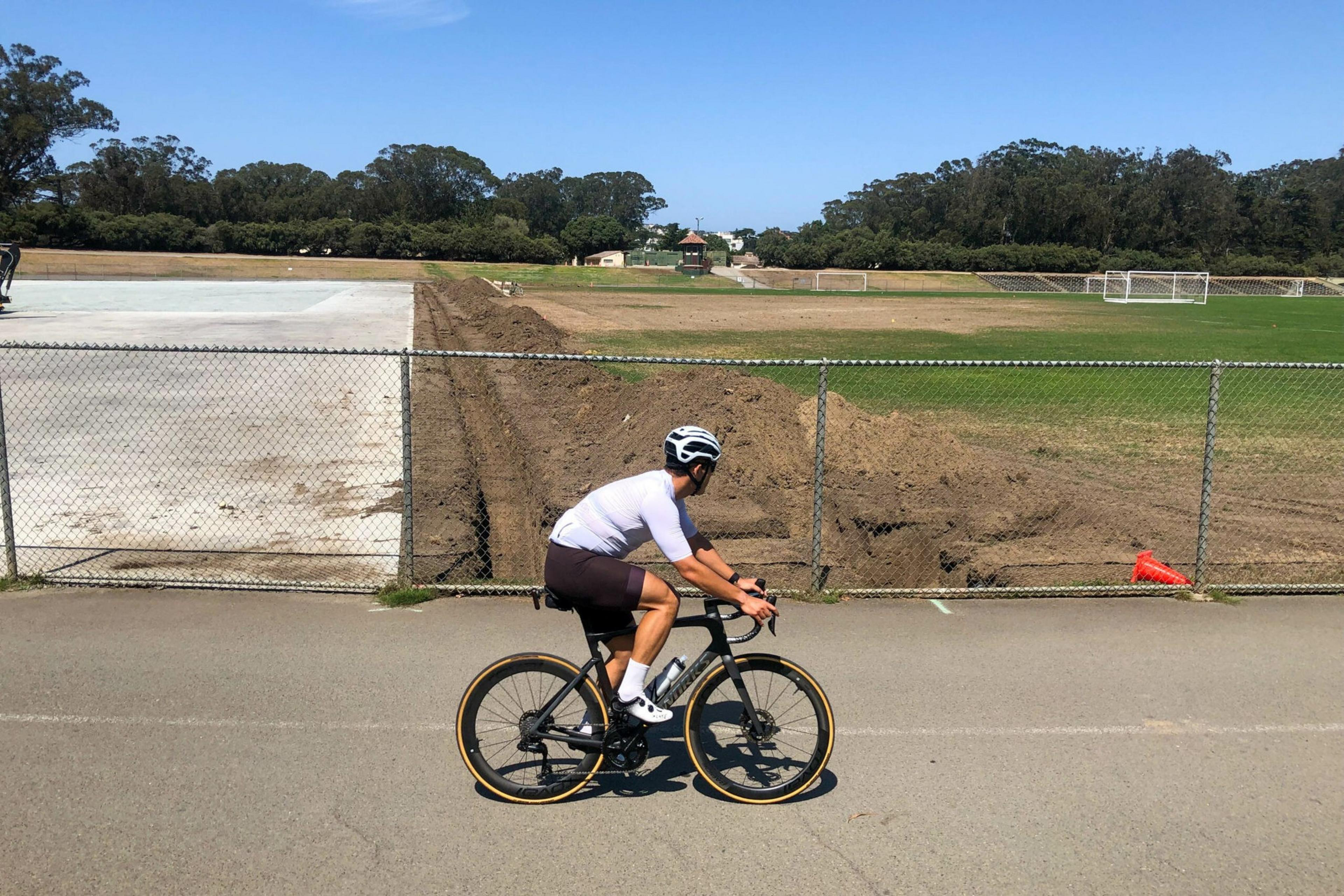 A cyclist wearing a white helmet and jersey rides a black bike on a paved path beside a field under a clear blue sky.