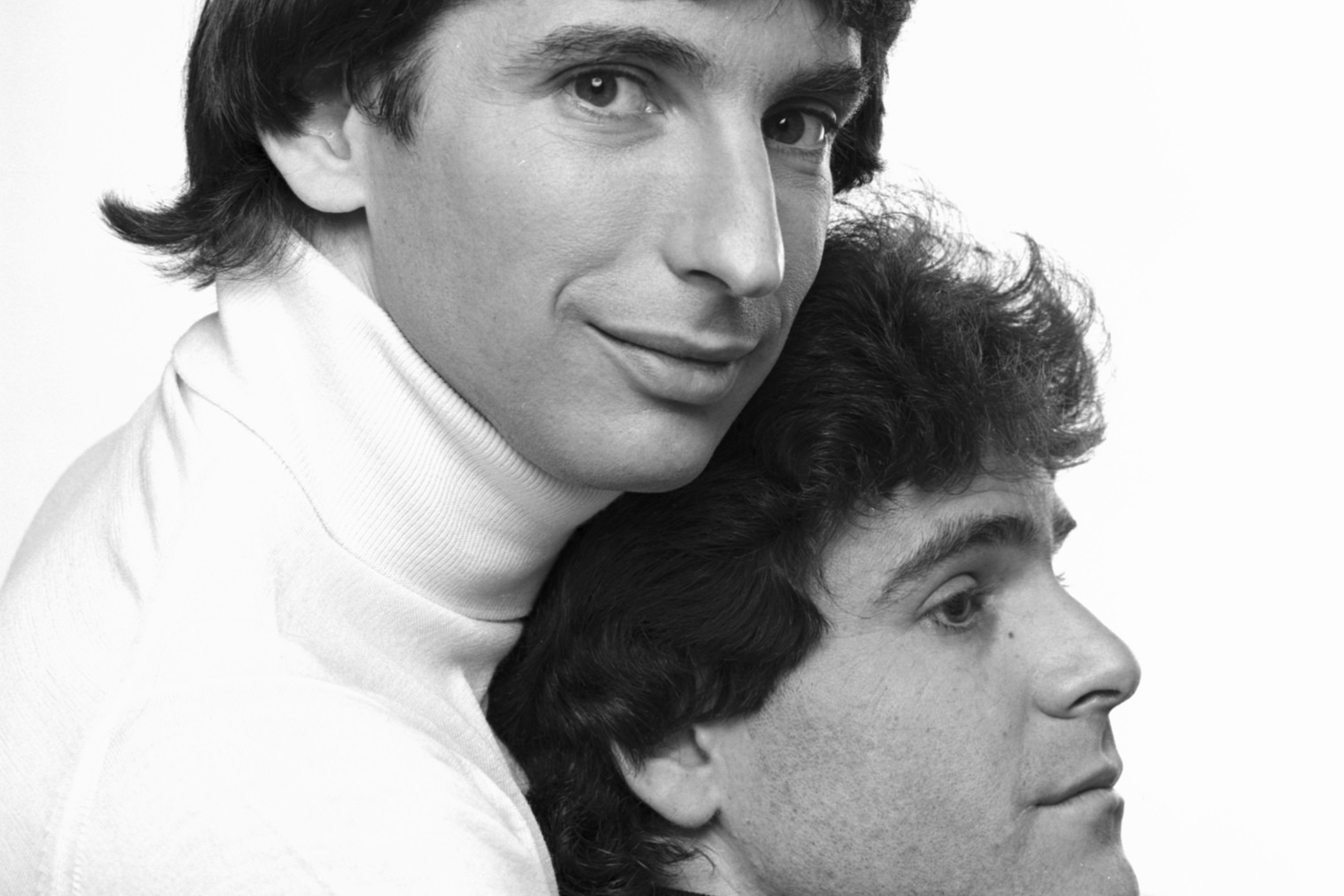 A man in a white turtleneck hugs from behind a seated man in a black turtleneck, with both facing opposite directions in a black-and-white photo.