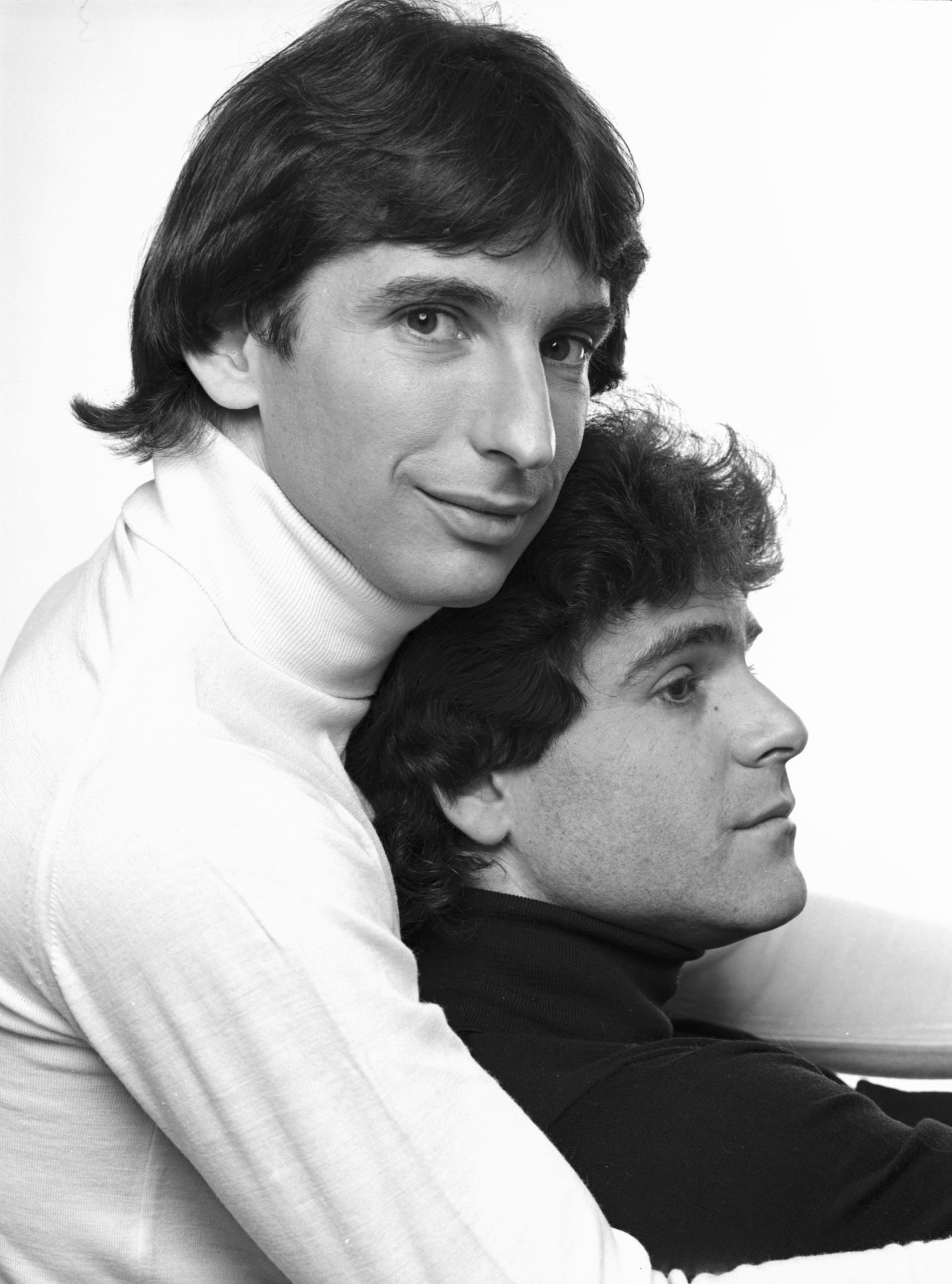 A man in a white turtleneck hugs from behind a seated man in a black turtleneck, with both facing opposite directions in a black-and-white photo.