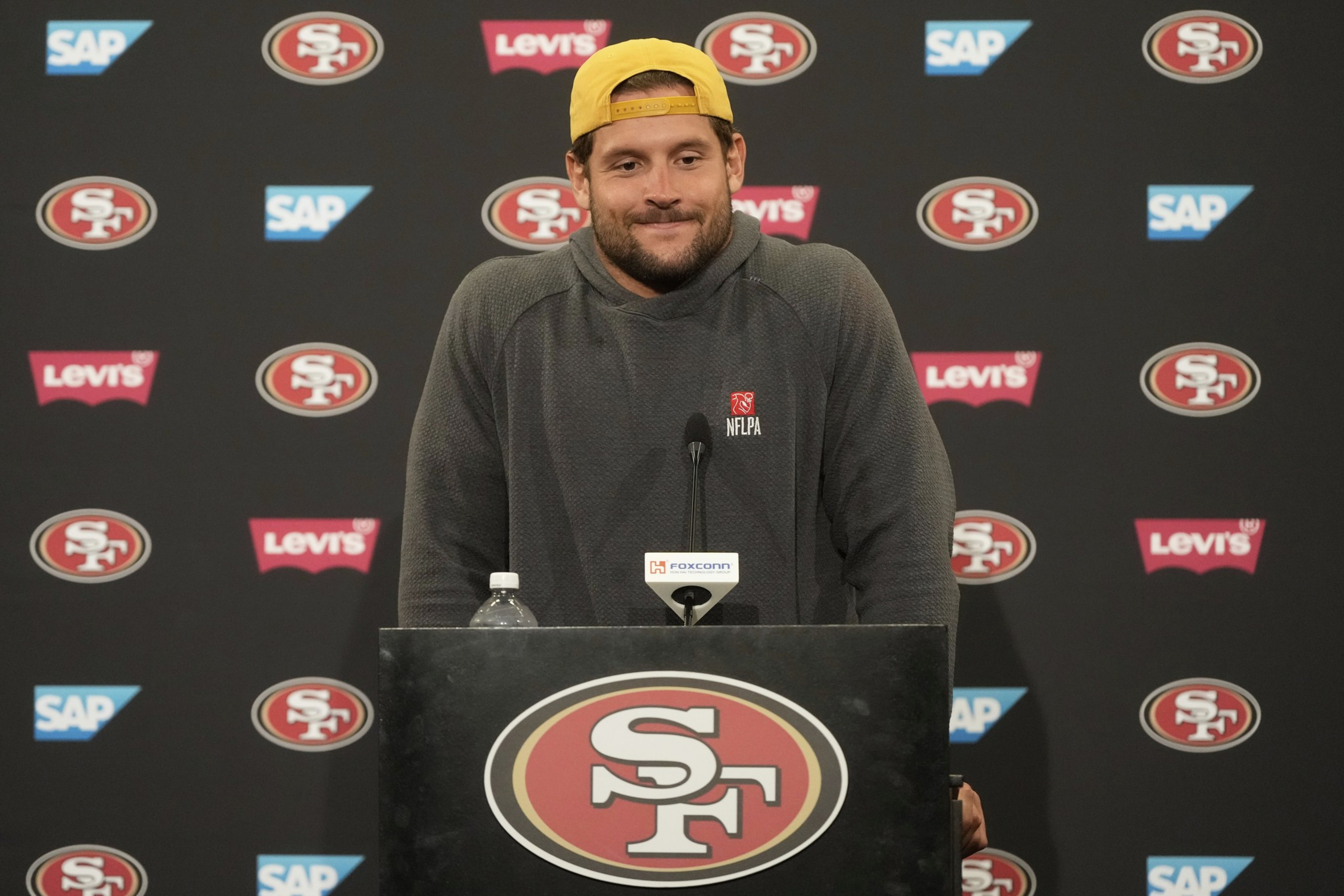 A person wearing a yellow cap and gray sweatshirt stands at a podium with a 49ers logo, surrounded by SAP and Levi's logos on a black backdrop.