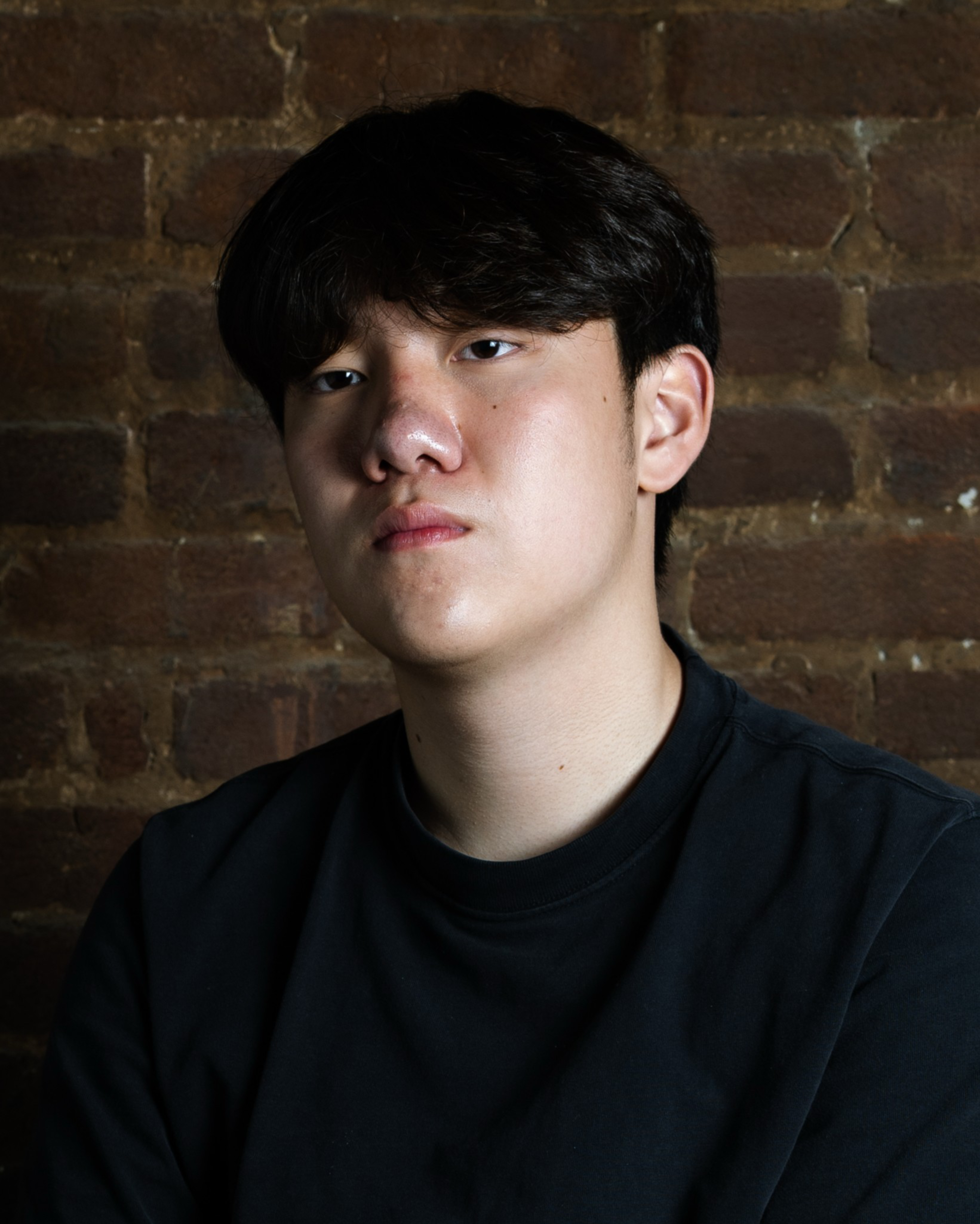 A young man with short black hair and light skin wears a black shirt, looking directly at the camera with a neutral expression against a brick wall.