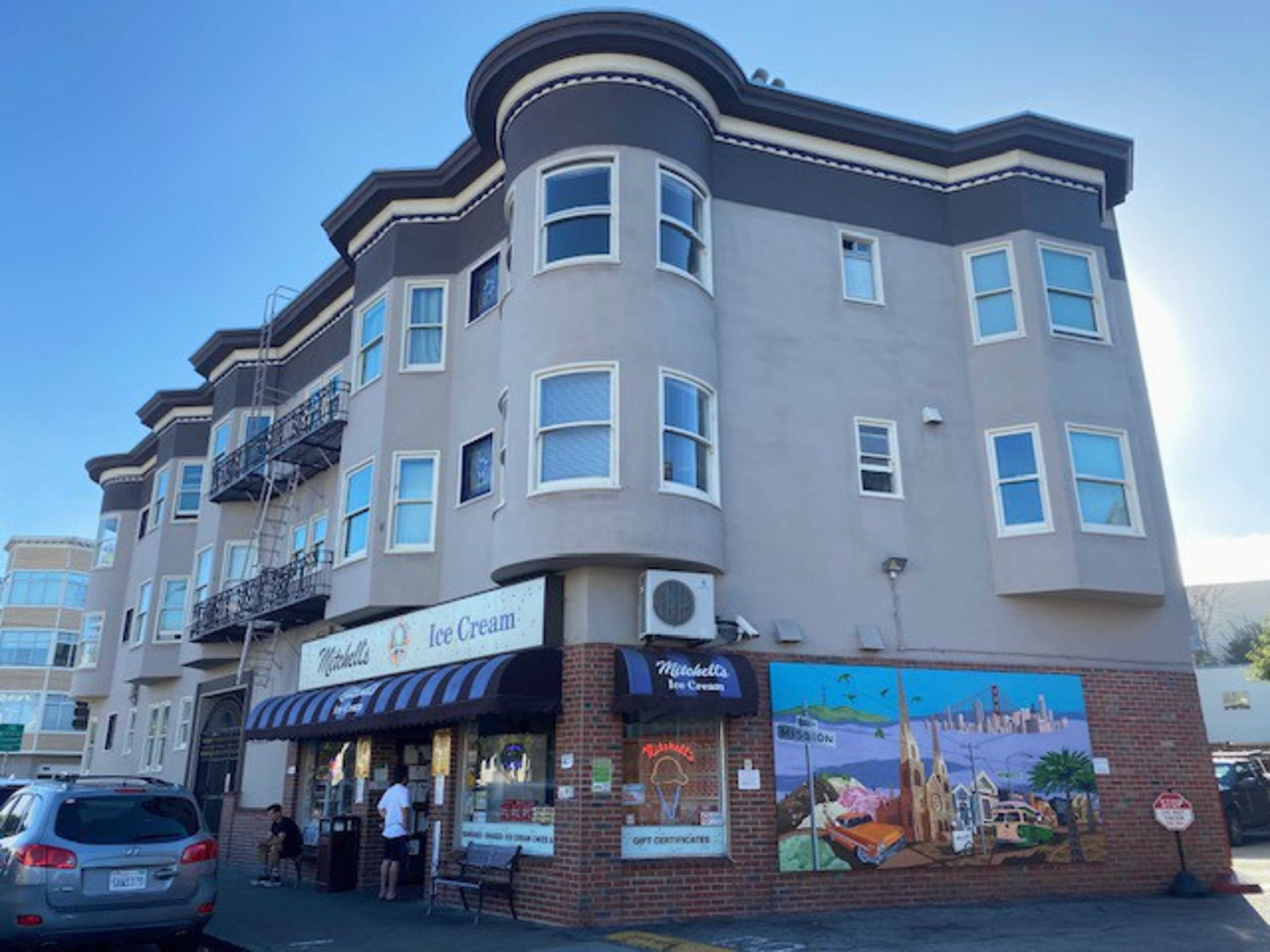 The image shows a beige, multi-story building with an ice cream shop on the ground floor. The shop has a sign and a striped awning, with a mural painted on the side wall.