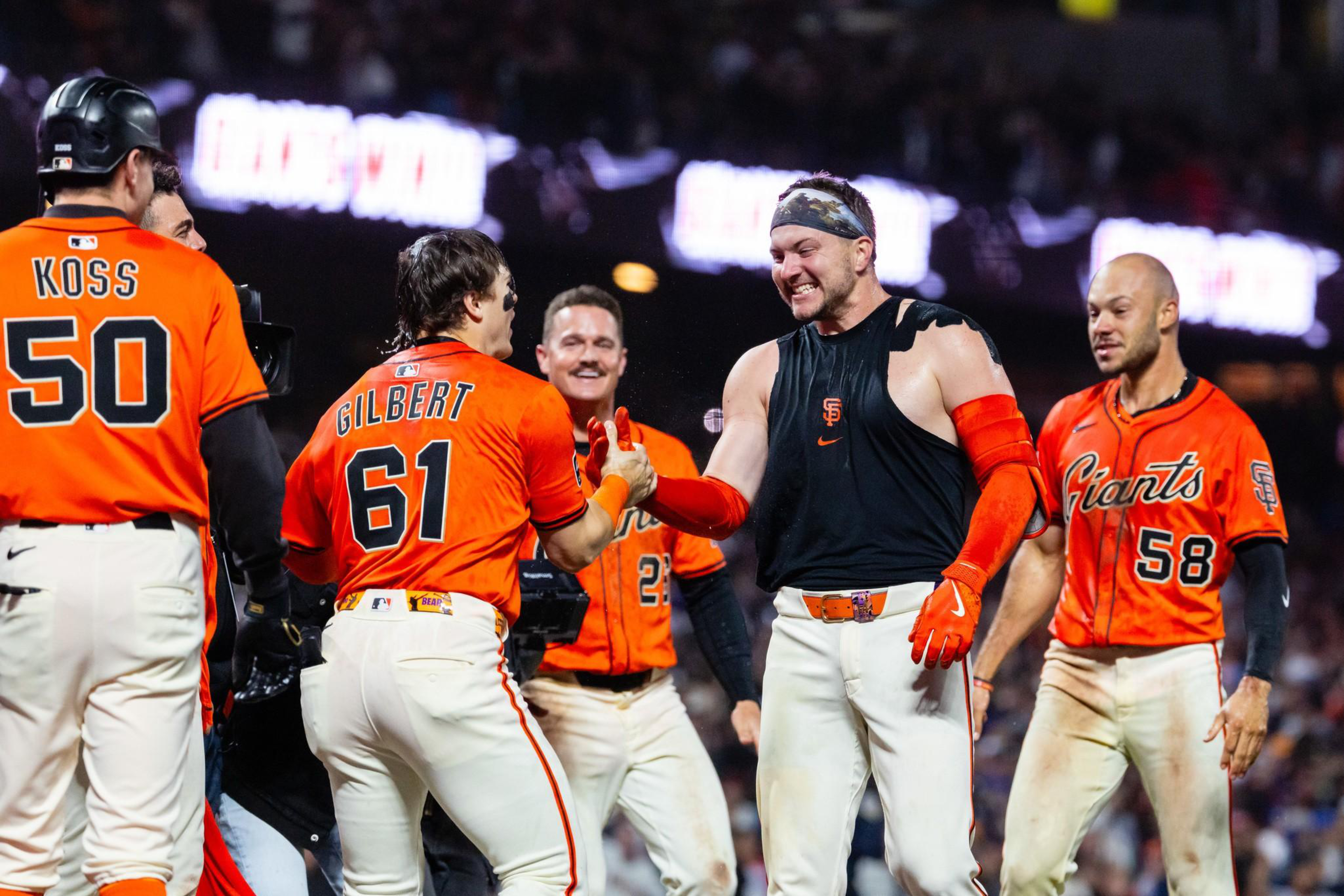 San Francisco Giants baseball players in orange jerseys celebrate on the field, with one player in sleeveless black congratulating a teammate.