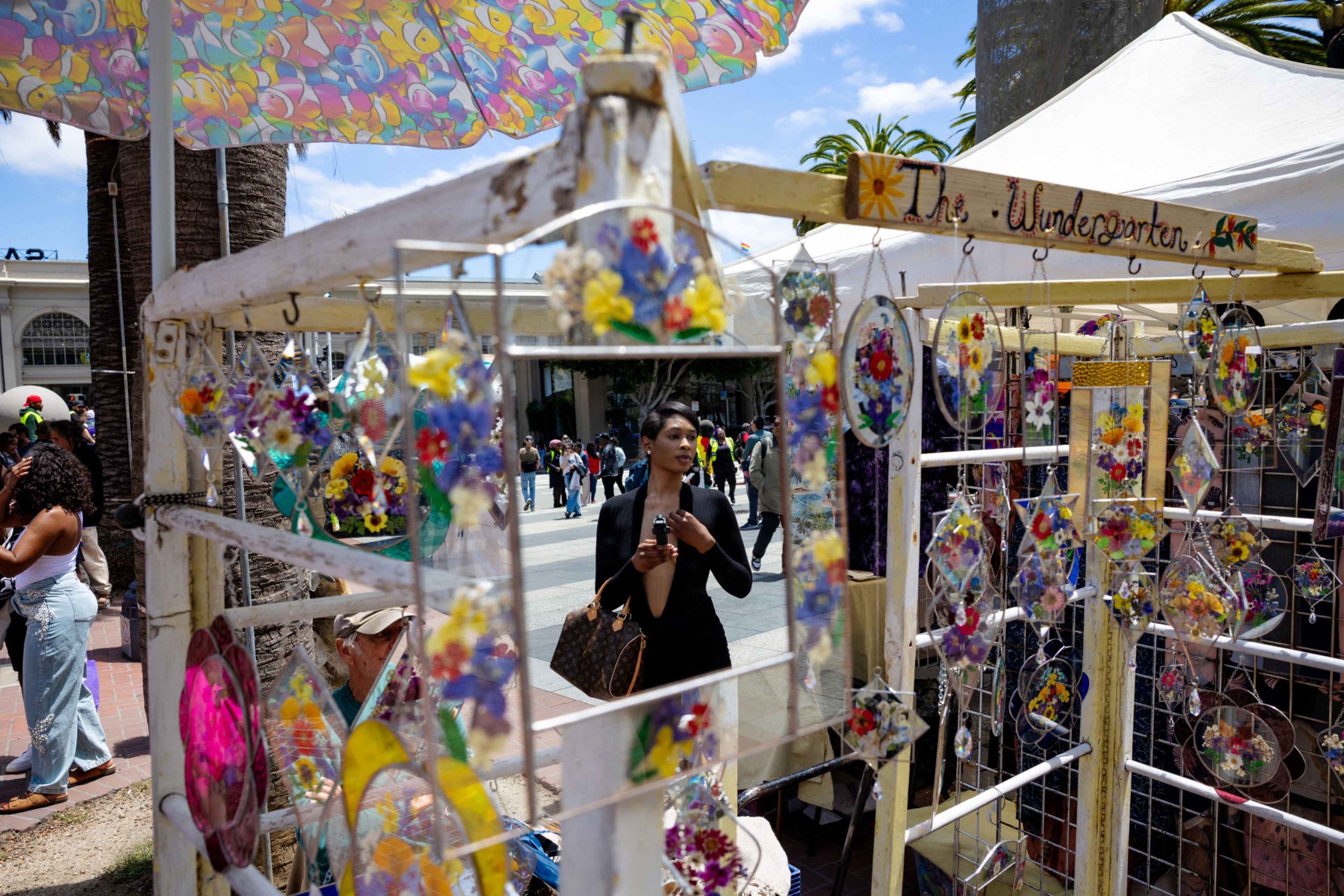 A market stall displays colorful stained-glass floral decorations and mirrors, with a woman reflected in one mirror and people walking in the background.