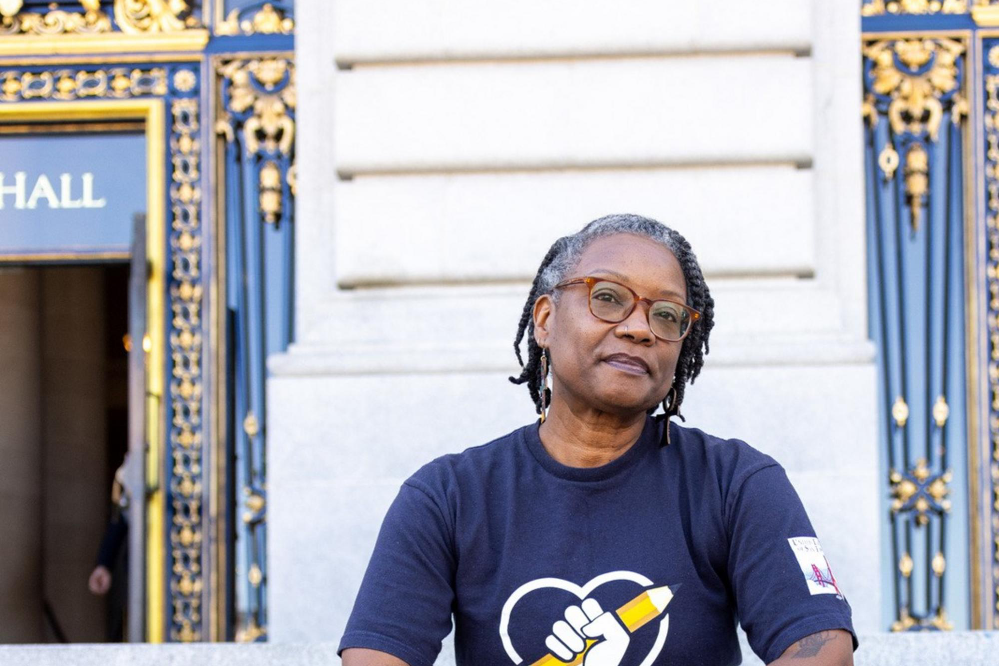 A person with dreadlocks, wearing glasses, a navy shirt with a fist and pencil logo, dark pants, and black Crocs, sits on ornate building steps with golden details.