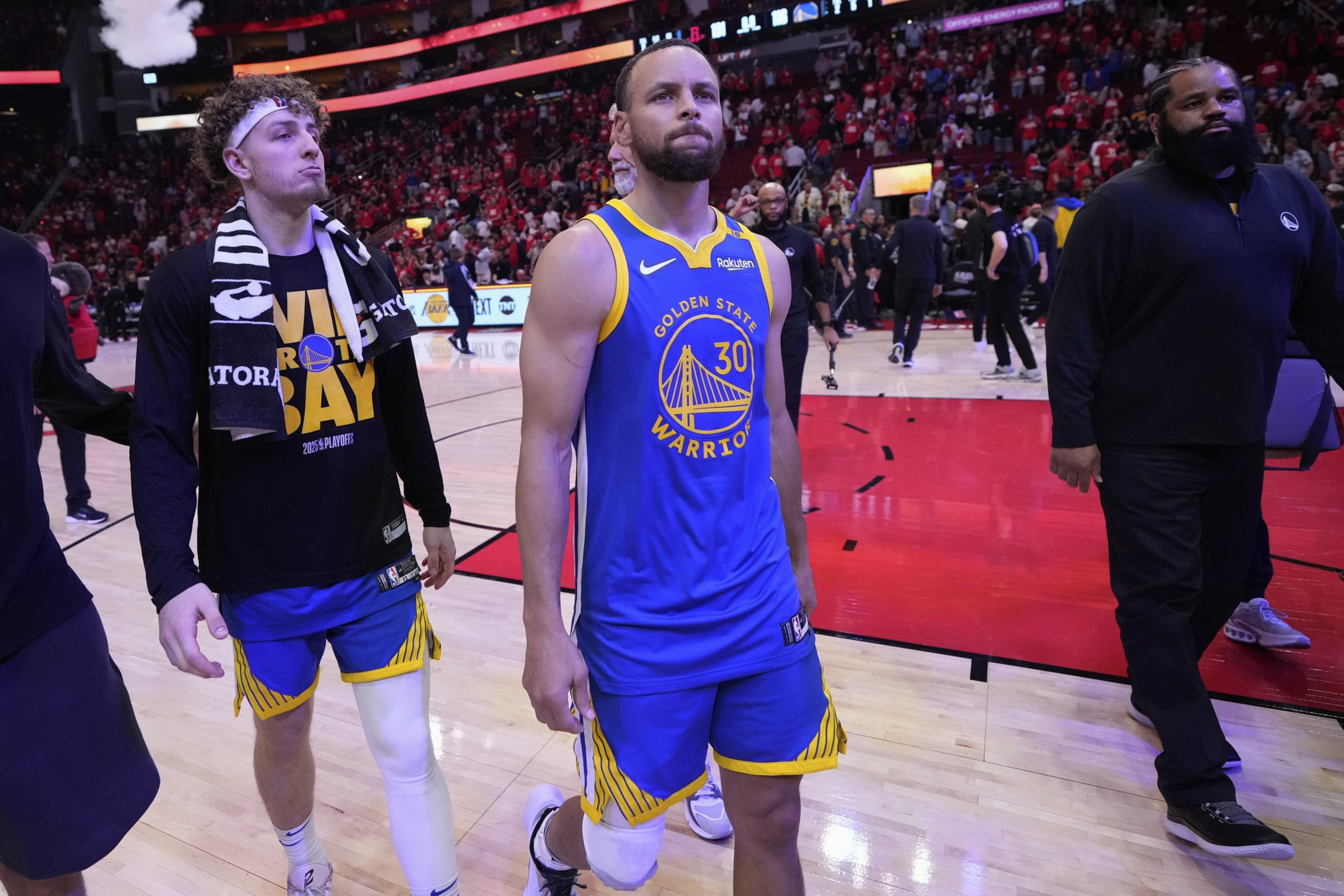 Two basketball players walk on the court, one in a blue Golden State Warriors jersey, the other in a team shirt with a towel over his shoulder, surrounded by a crowd.
