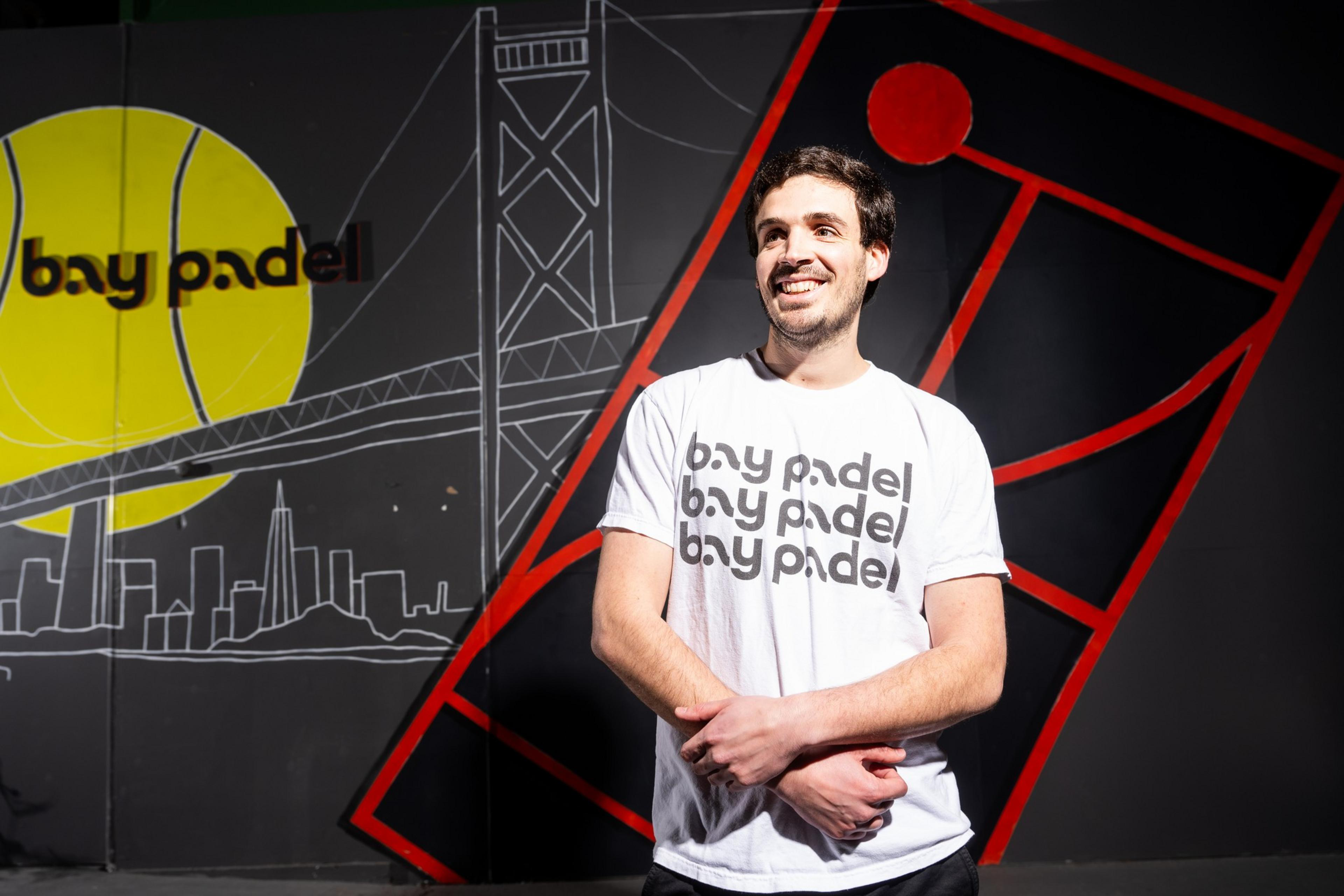 A person in a white &quot;bay padel&quot; T-shirt stands smiling against a mural with a bridge, city skyline, and abstract red and black geometric pattern.