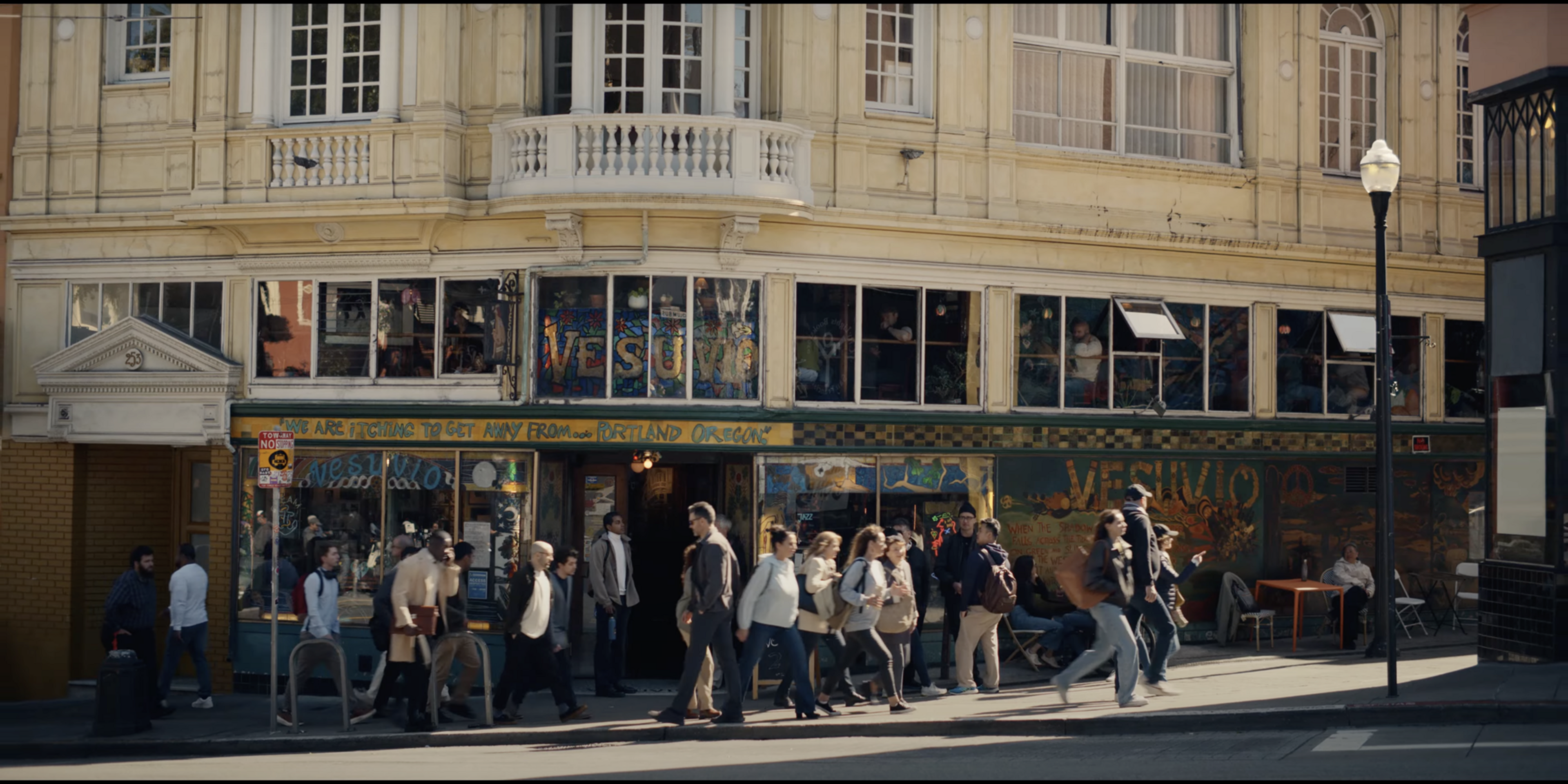 A busy street scene shows a group of people walking past a vintage-looking corner café named &quot;Vesuvio.&quot; The building features large windows and intricate architecture.