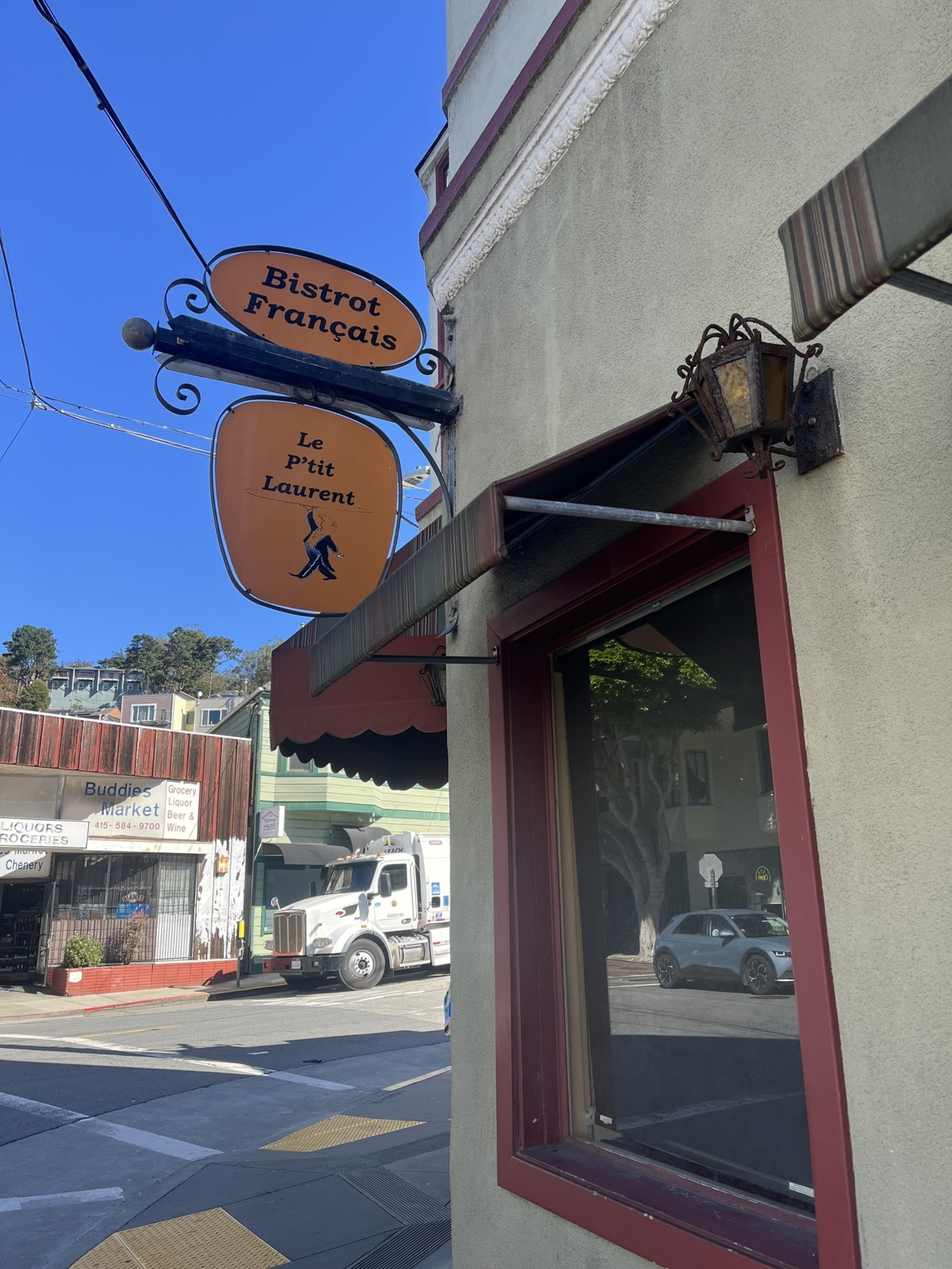 A French bistro sign reading “Bistrot Français Le p’tit Laurent” hangs above a window on a sunny street corner with vehicles and a market across the street.