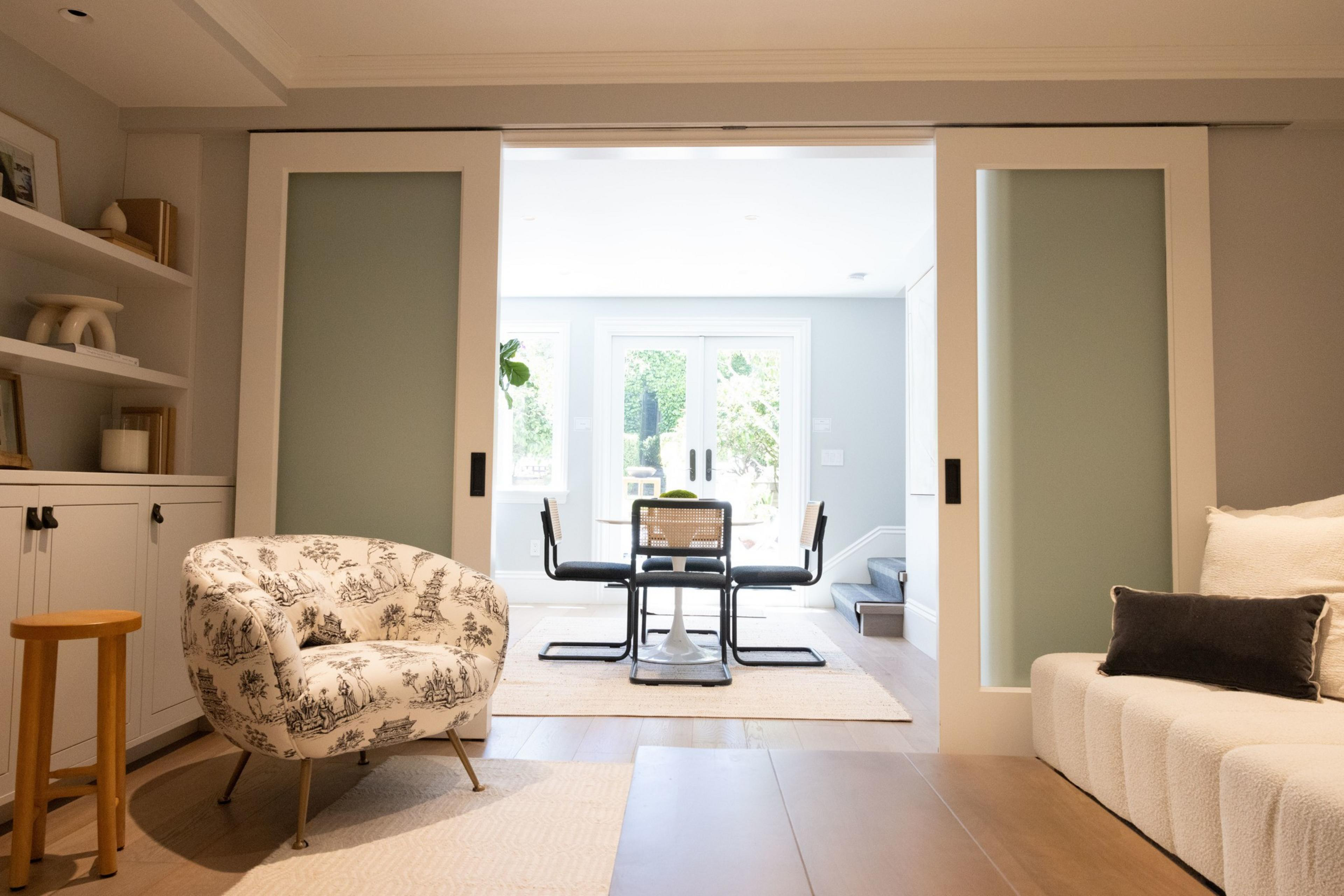 A cozy living room features a floral armchair, a cushioned sofa, and a wooden stool. Sliding doors open to a dining area with a modern table, chairs, and a bright window view.