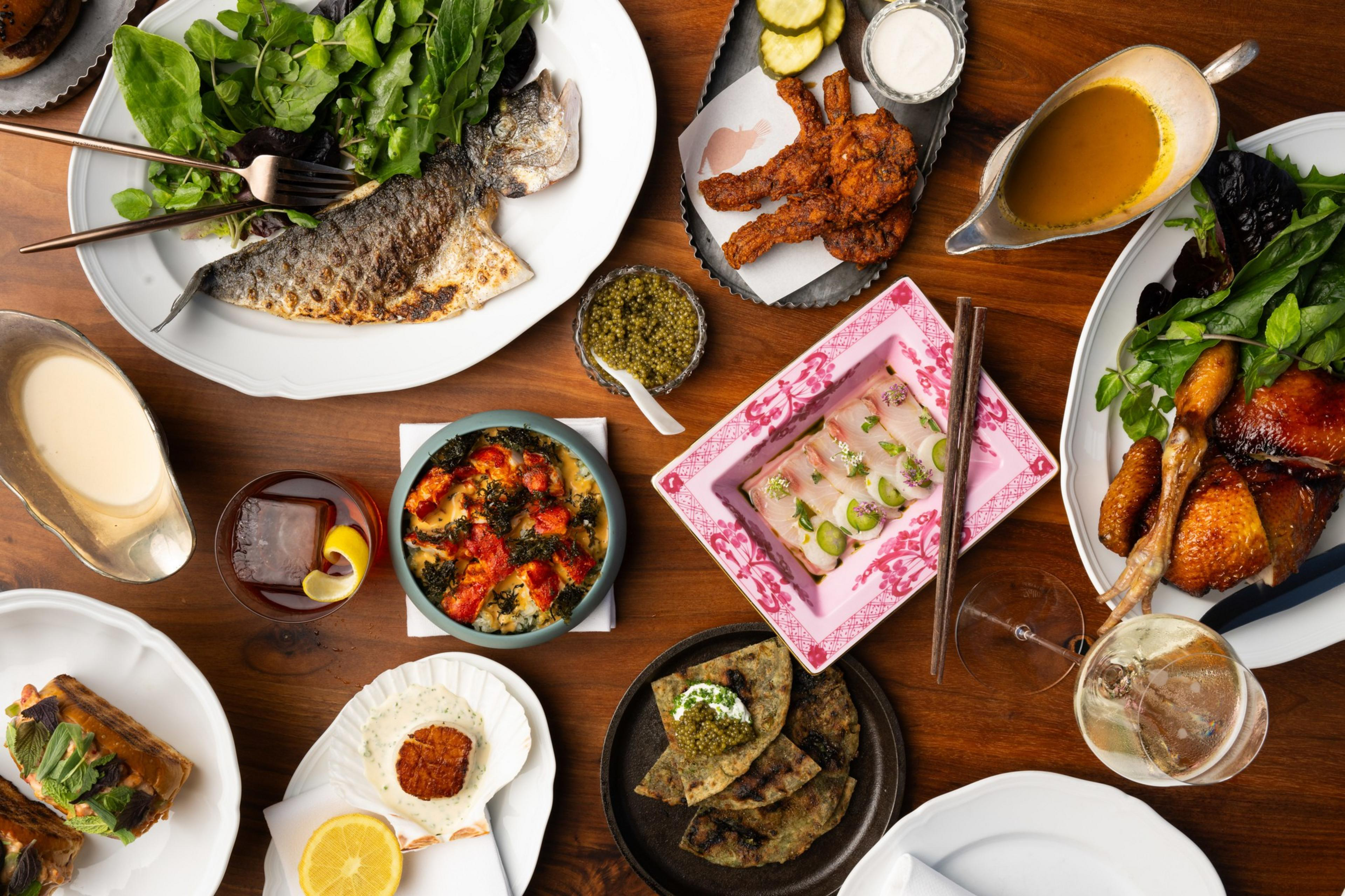 A variety of dishes are displayed on a wooden table including fish, fried chicken, salad, a cocktail, sauces, and garnished vegetables.