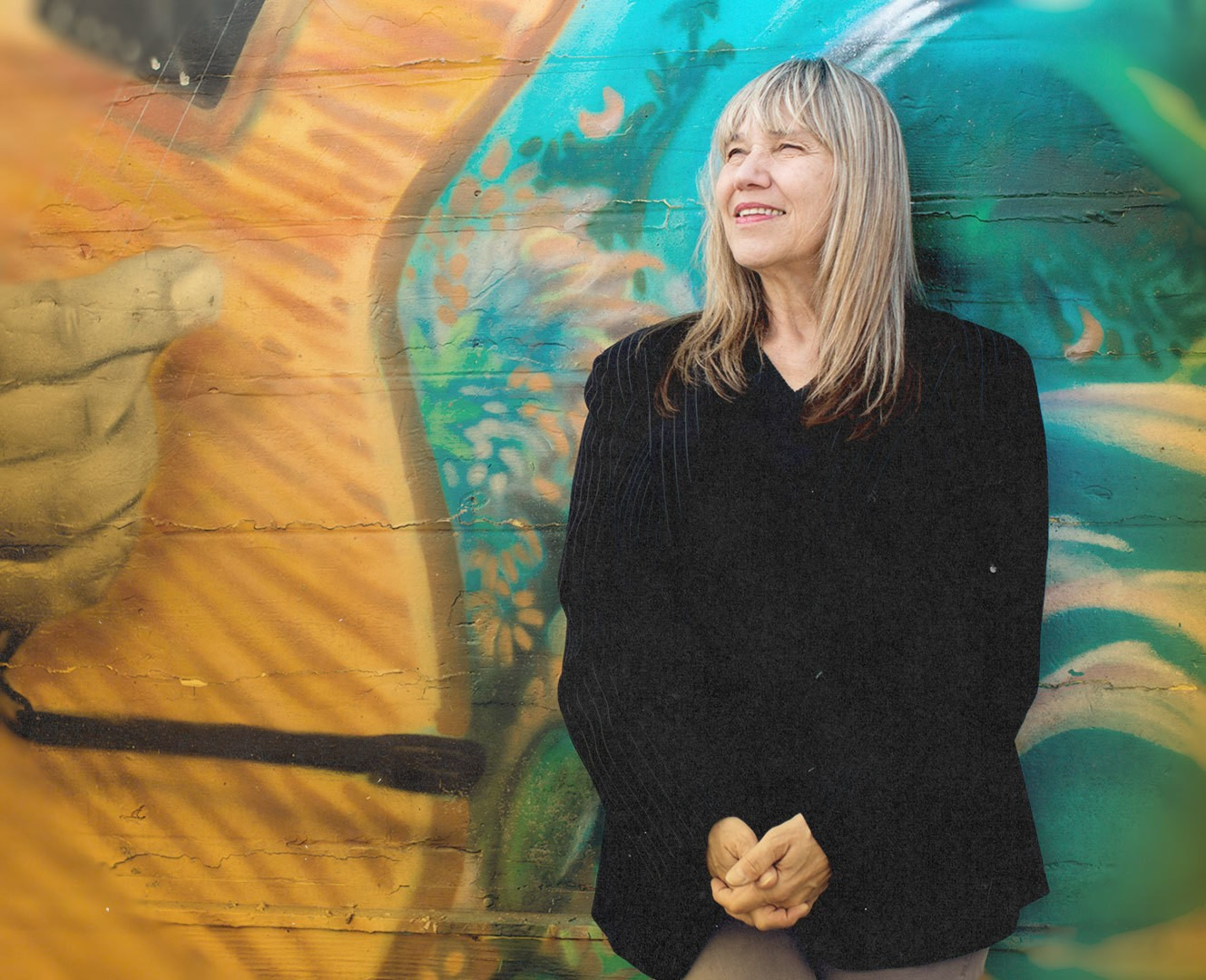 A woman in a black suit stands smiling in front of a mural in the Mission.