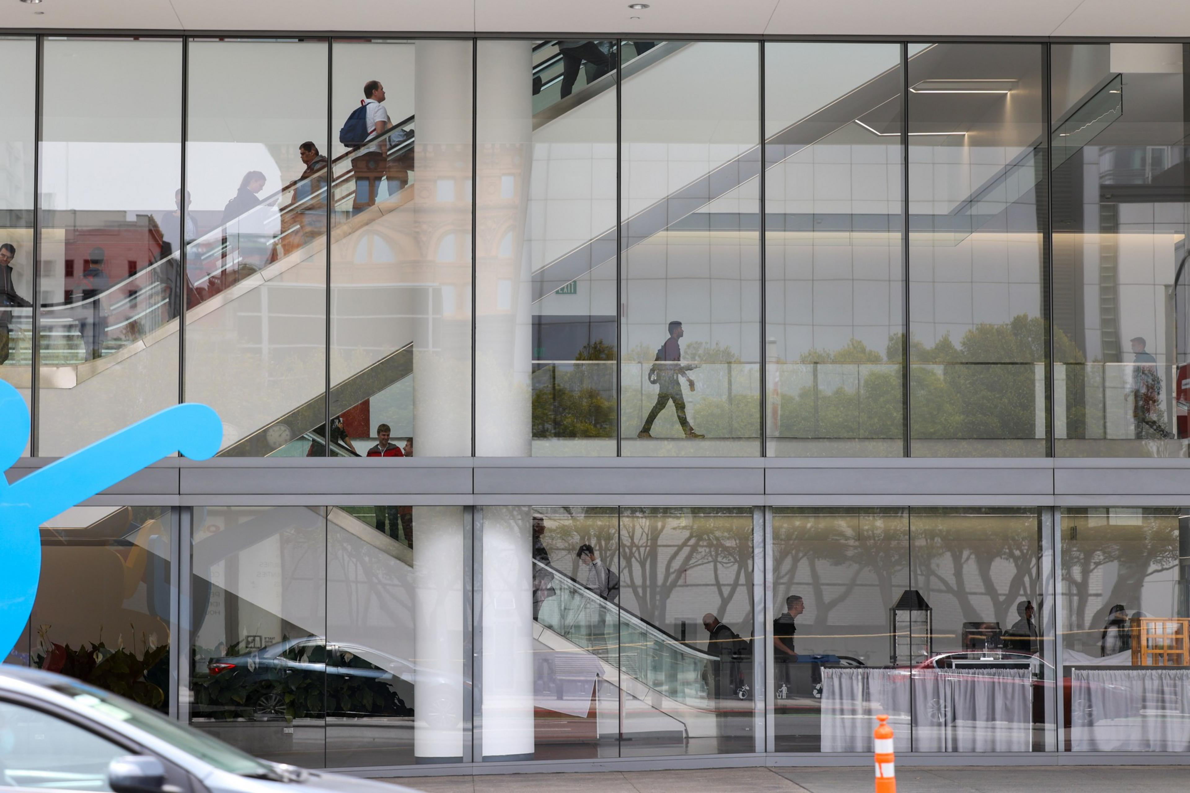 People are walking inside a glass building with escalators visible on two floors, reflecting outdoor surroundings and vehicles on the street.