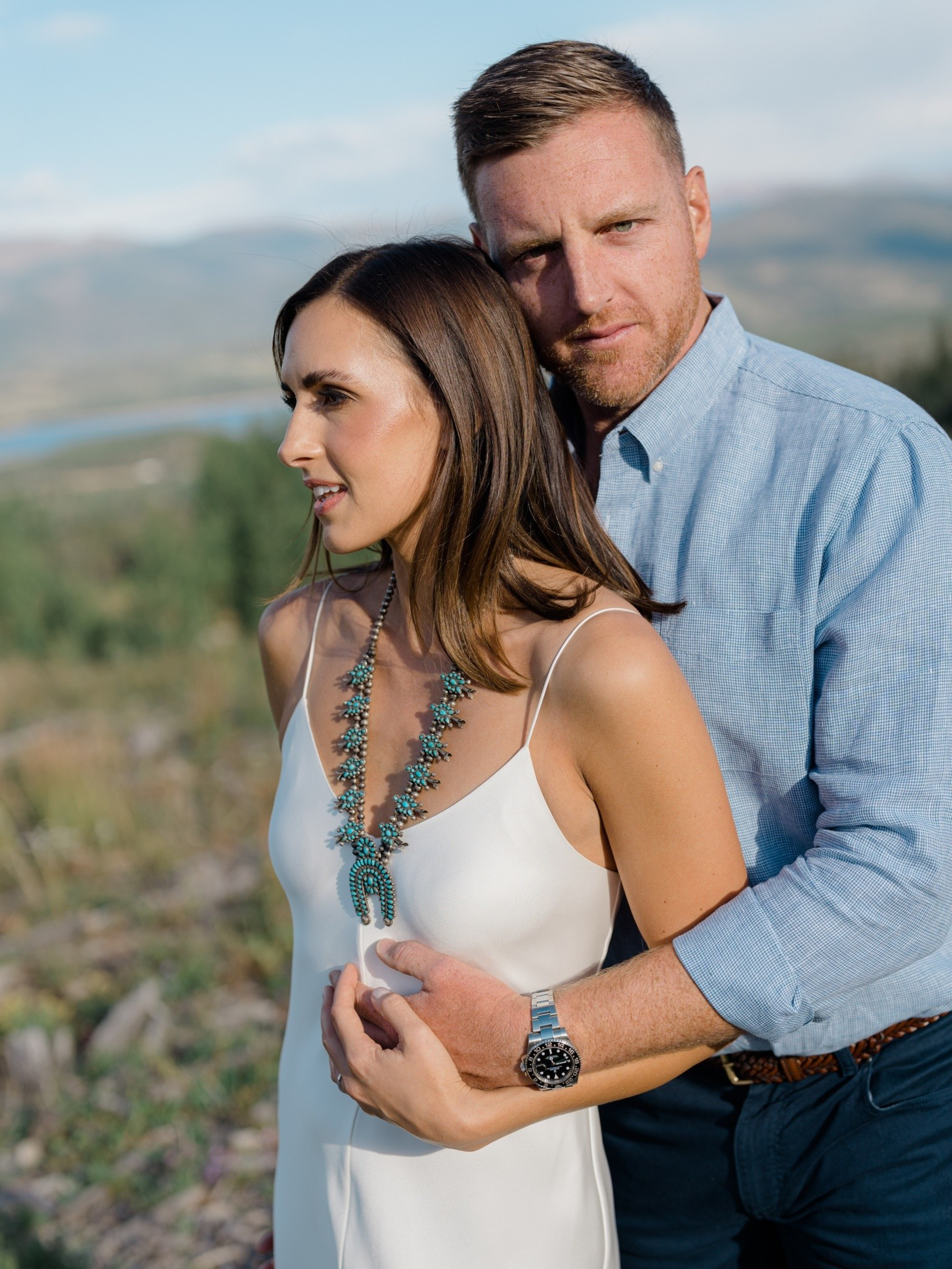A couple stands embracing outdoors. The woman wears a white dress with turquoise jewelry, while the man is in a blue shirt. The background is a scenic landscape.