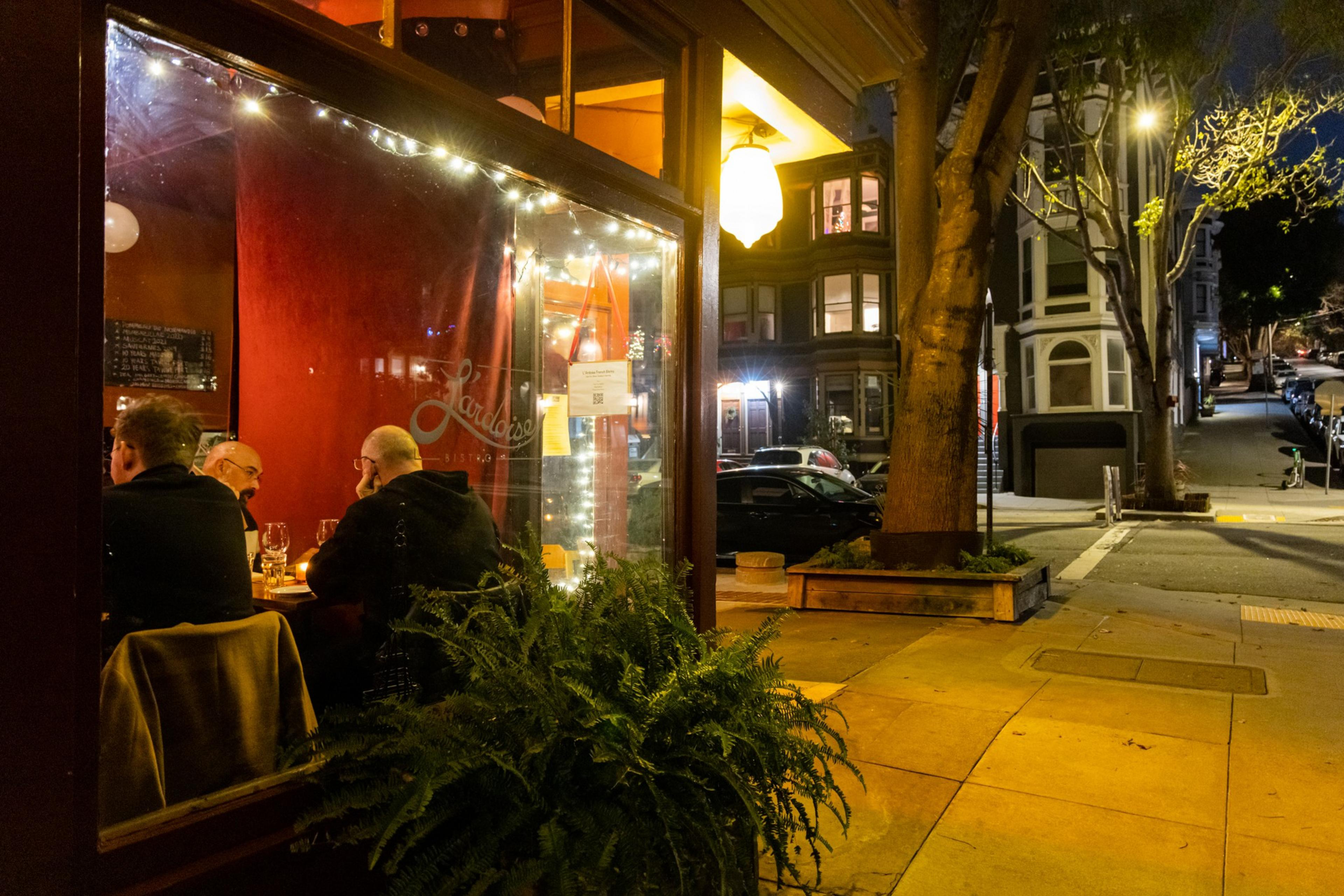 A cozy bistro corner with warm, glowing lights shows three people dining inside near a window. Outside, it's nighttime with a quiet street, trees, and parked cars.