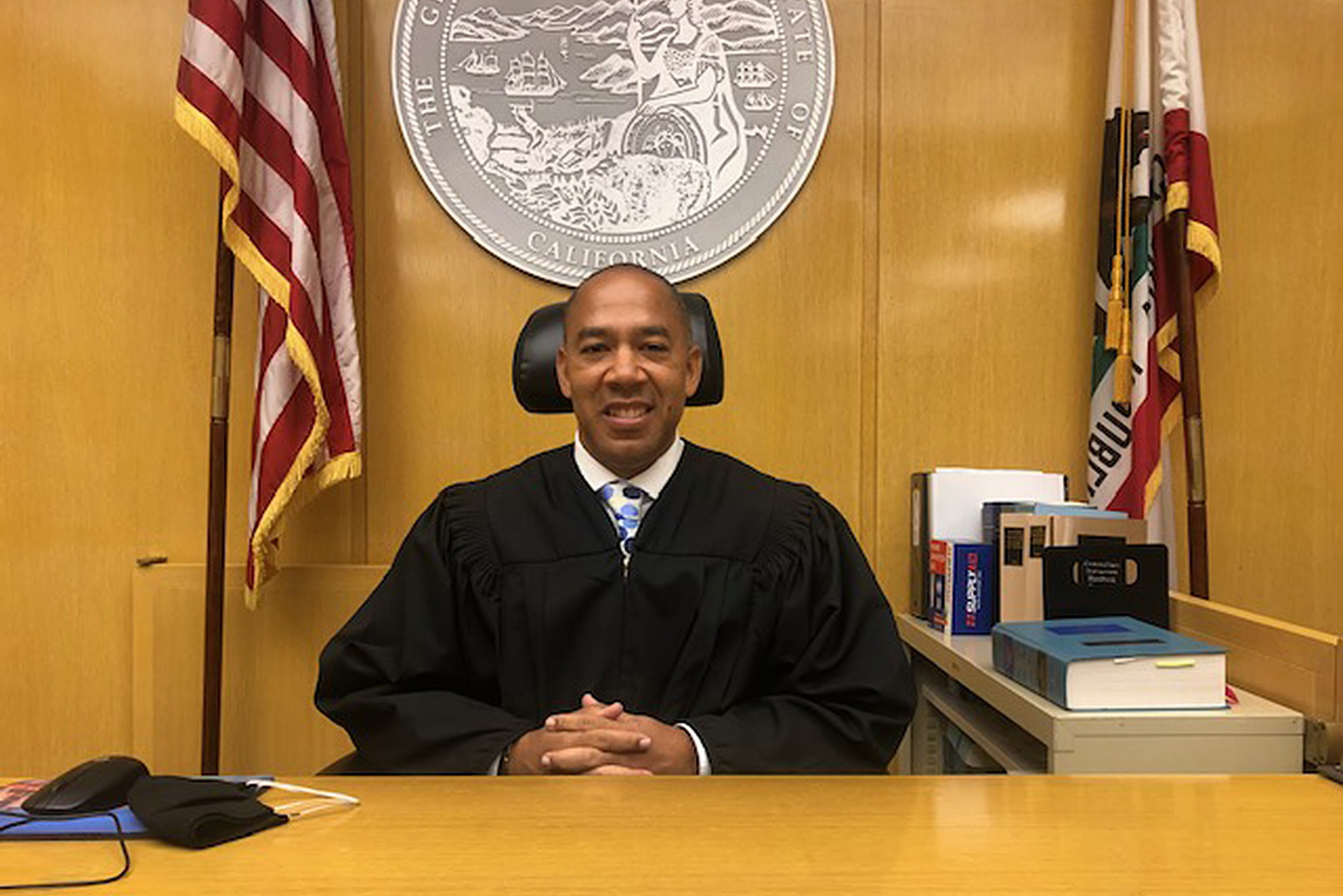 A smiling judge in a black robe sits at a wooden bench with folded hands, flanked by American and California state flags and legal books.
