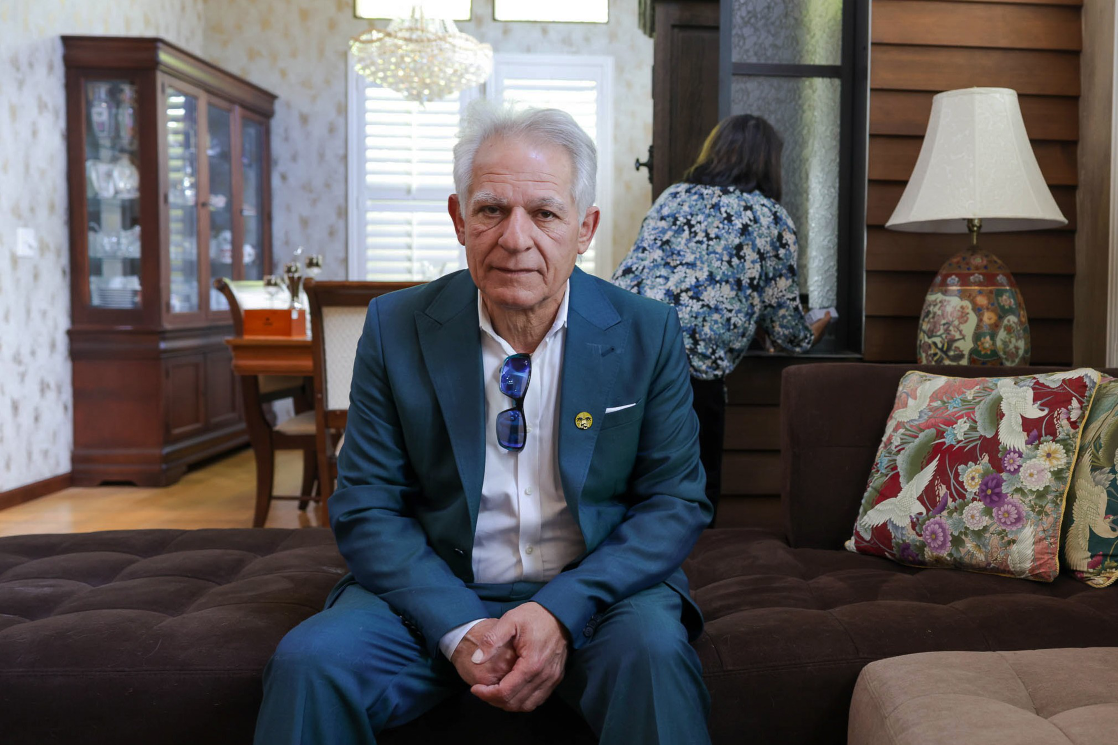 An elderly man in a blue suit sits on a brown ottoman; a woman looks out a window in a cozy room.