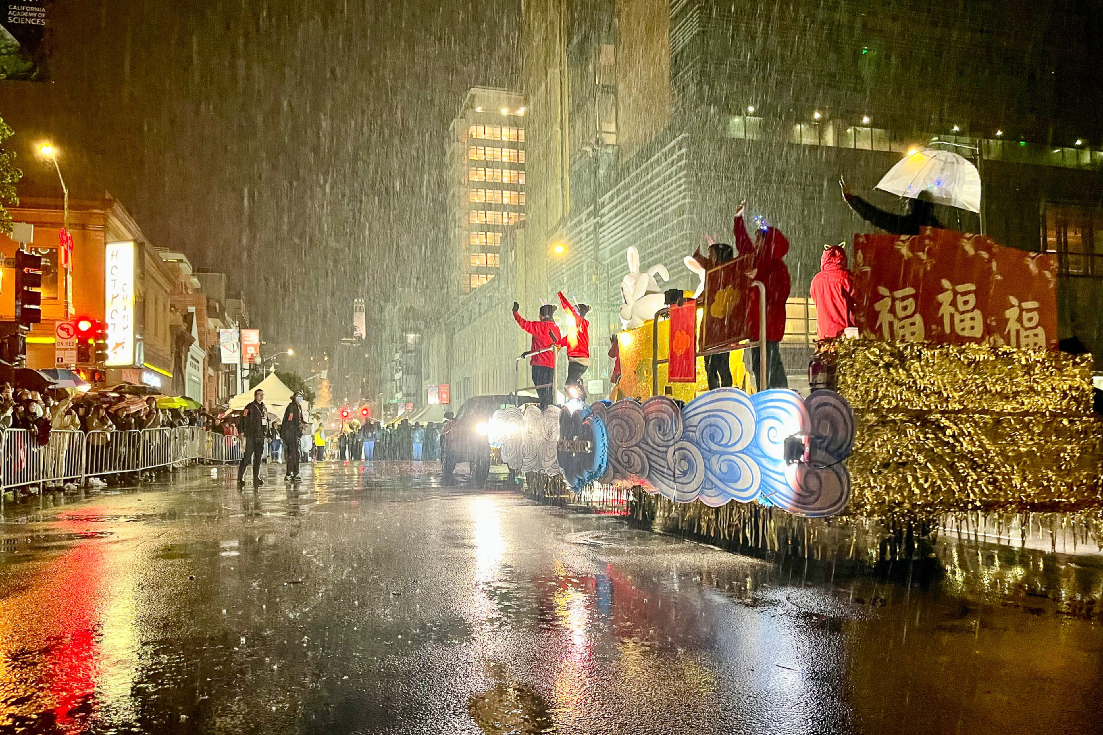 People in red costumes wave from a gold and blue lantern-adorned float during a rainy night parade with spectators behind barriers.