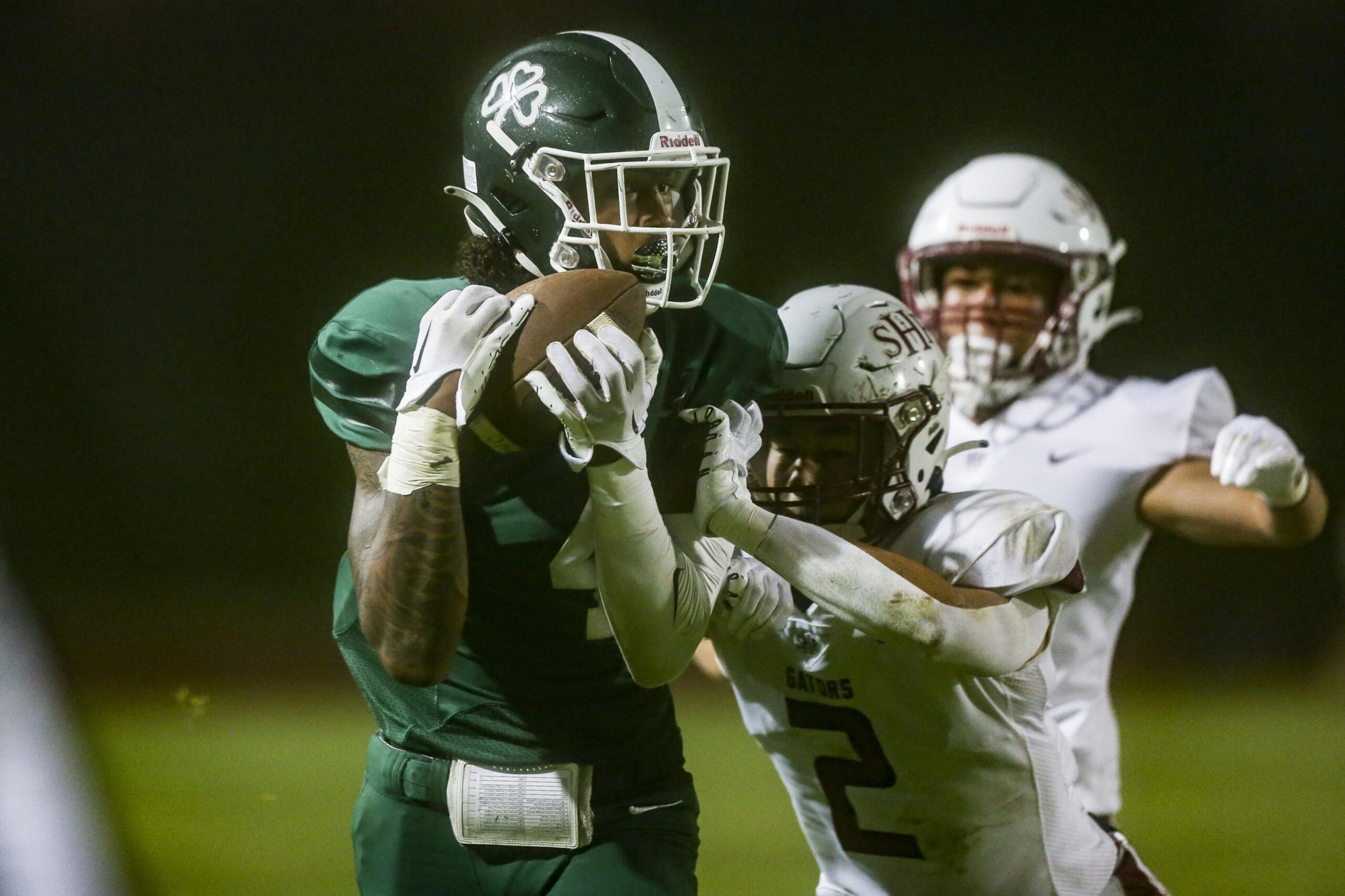 A football player in a green uniform tightly holds the ball while a player in white attempts to tackle him from the side during a night game.