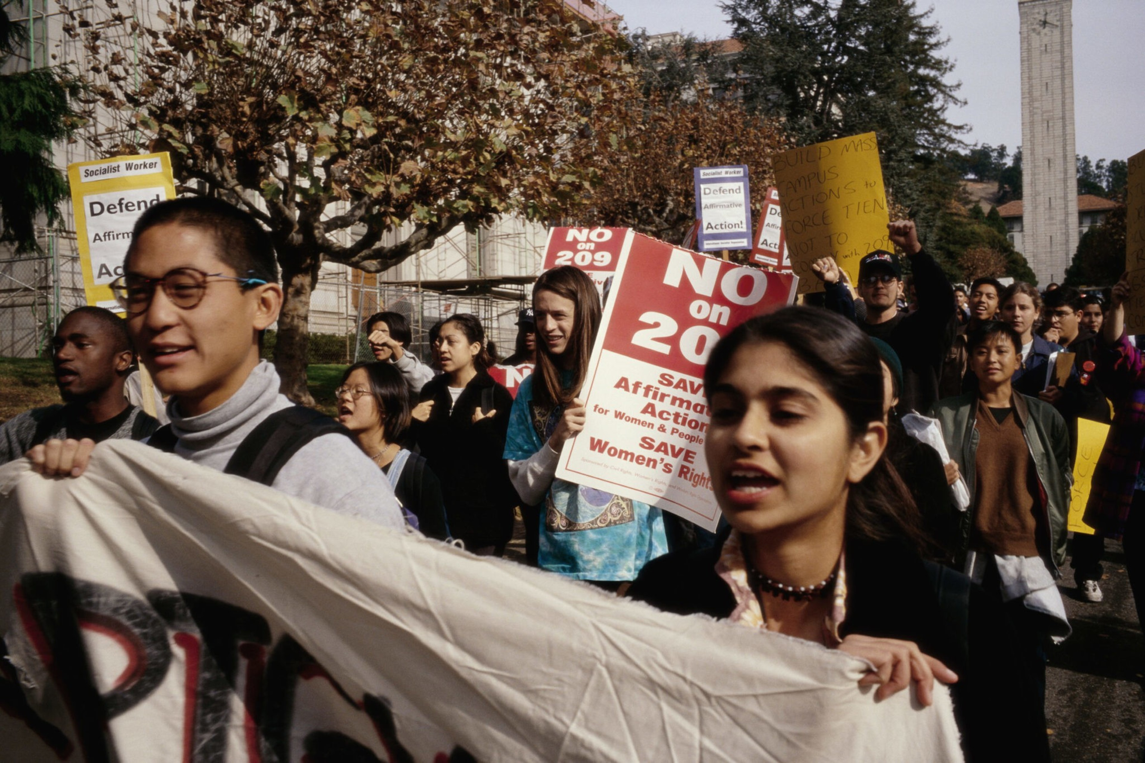 A diverse group of protesters march holding signs opposing "209" and advocating for women's rights and affirmative action.